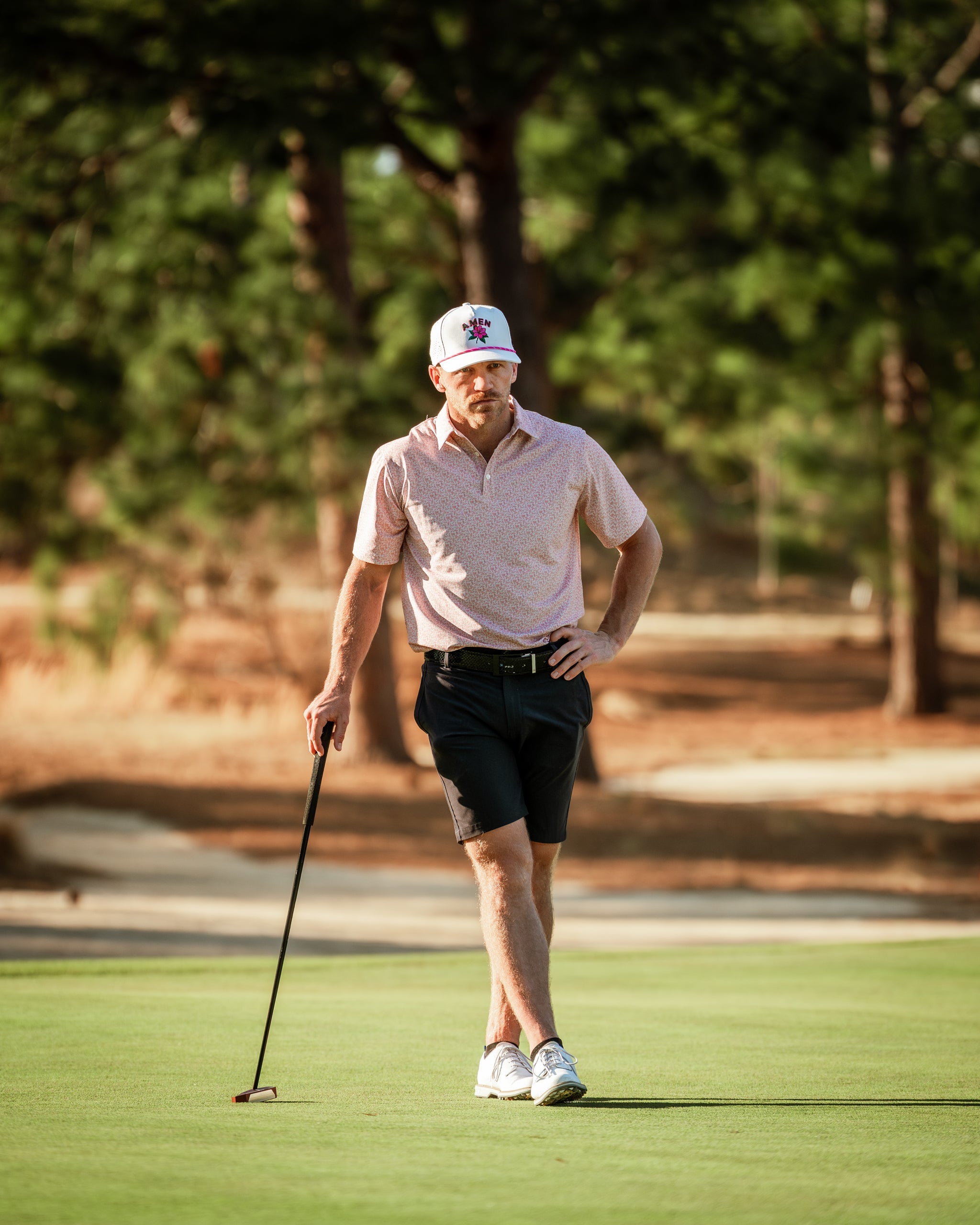 A golfer in a pink patterned polo and shorts walks on the green, holding a putter, surrounded by trees on a sunny day.