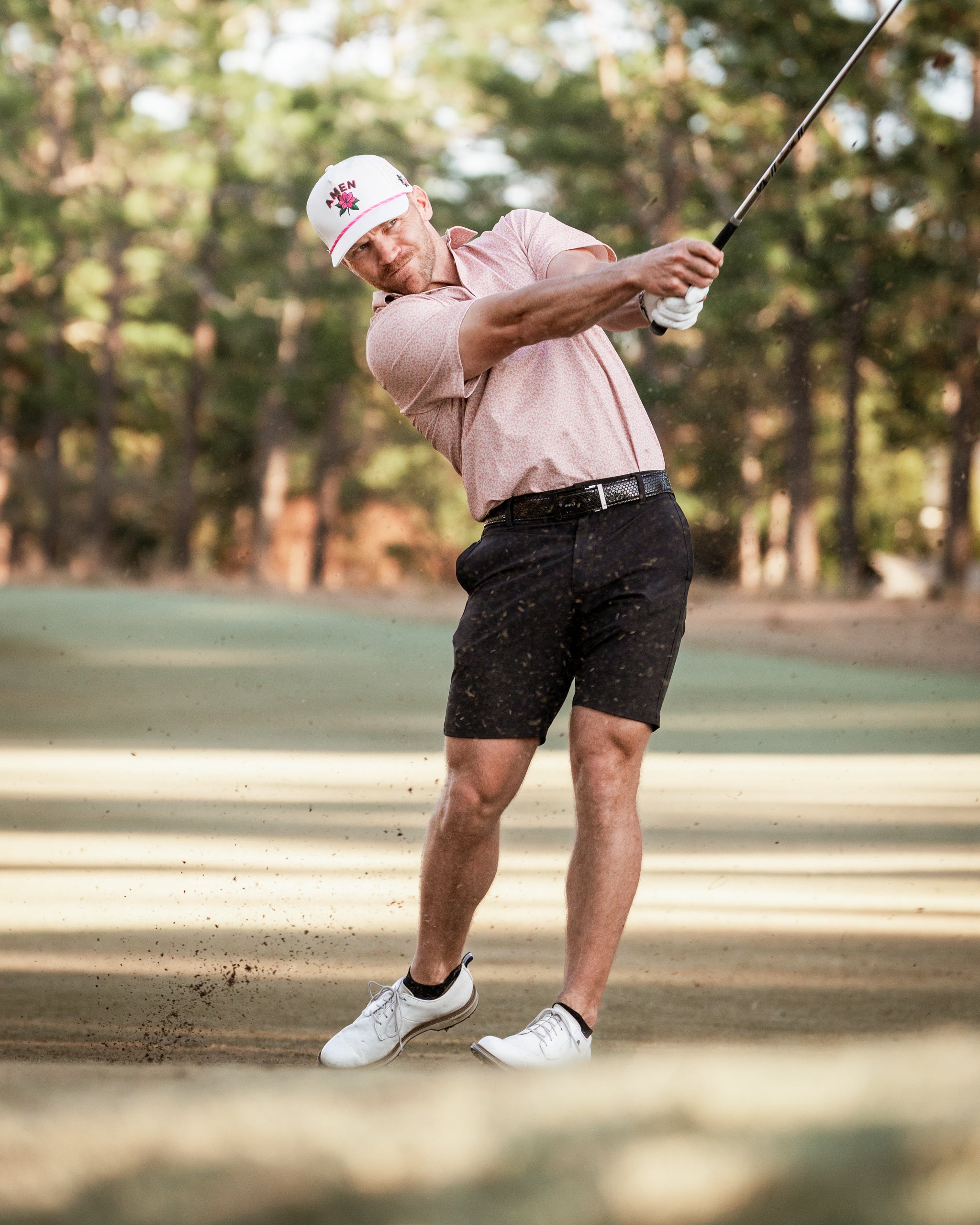 A golfer in a pink checkered shirt and black shorts swings a club on a sandy course, showcasing bold golf apparel.