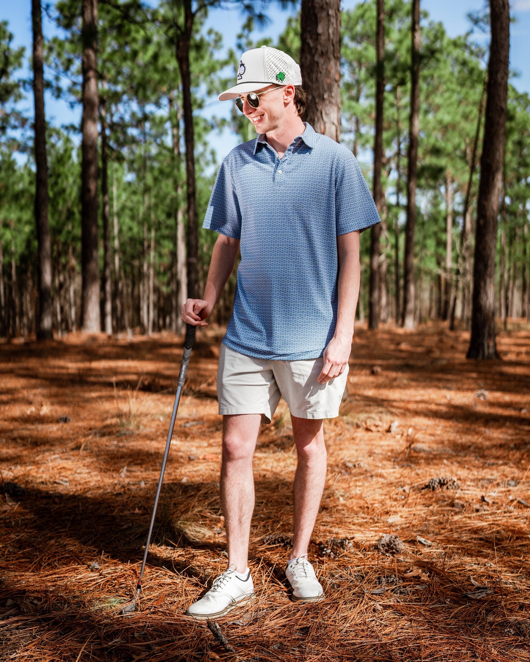 A golfer stands in a pine forest wearing a blue patterned polo shirt and beige shorts, holding a golf club.