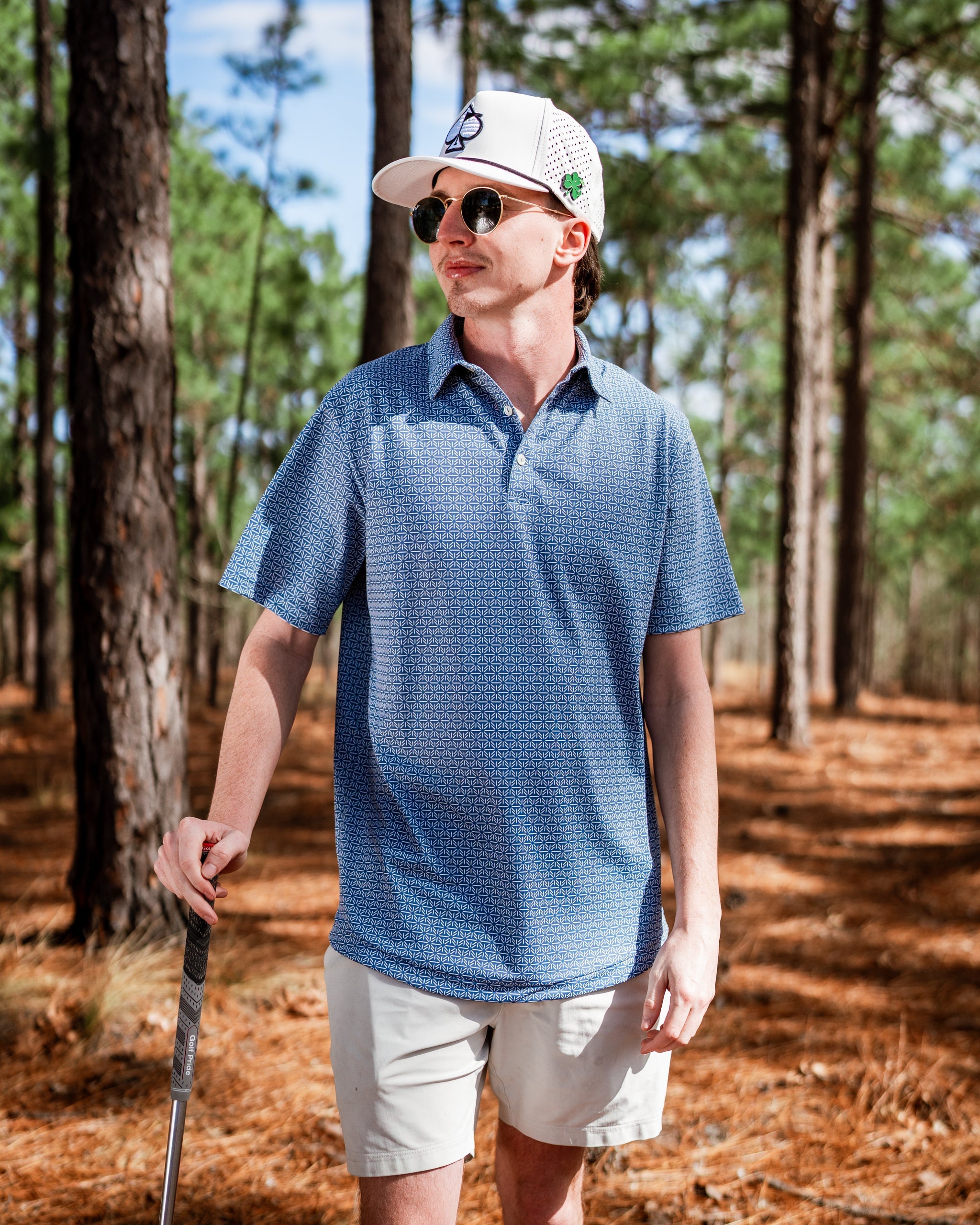 A golfer wearing a blue patterned polo shirt and a white cap stands in a pine forest, holding a golf club.