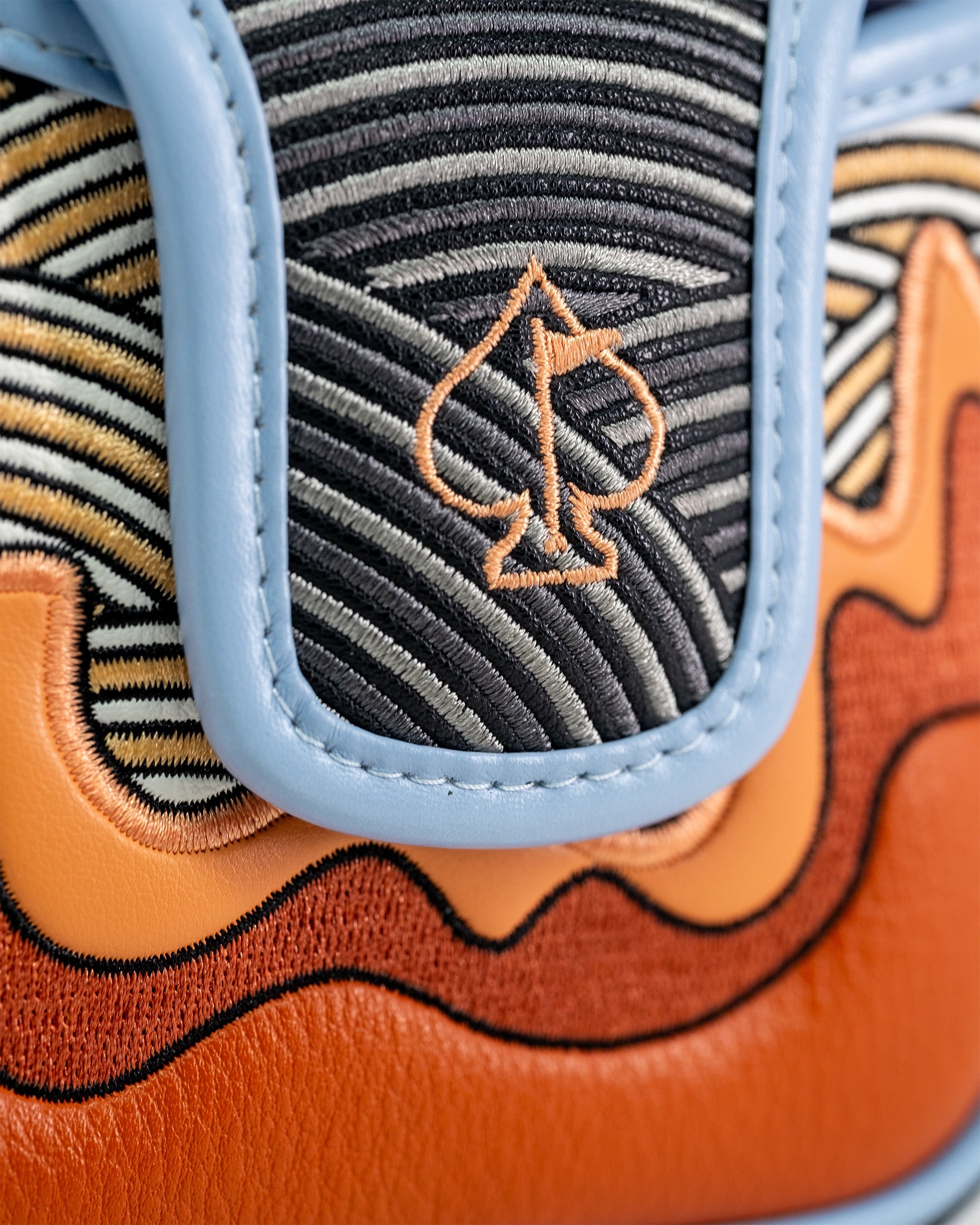Close-up of a golf mallet putter cover featuring embroidered black, orange, and gray striped fabric with a spade logo, edged in light blue leather.