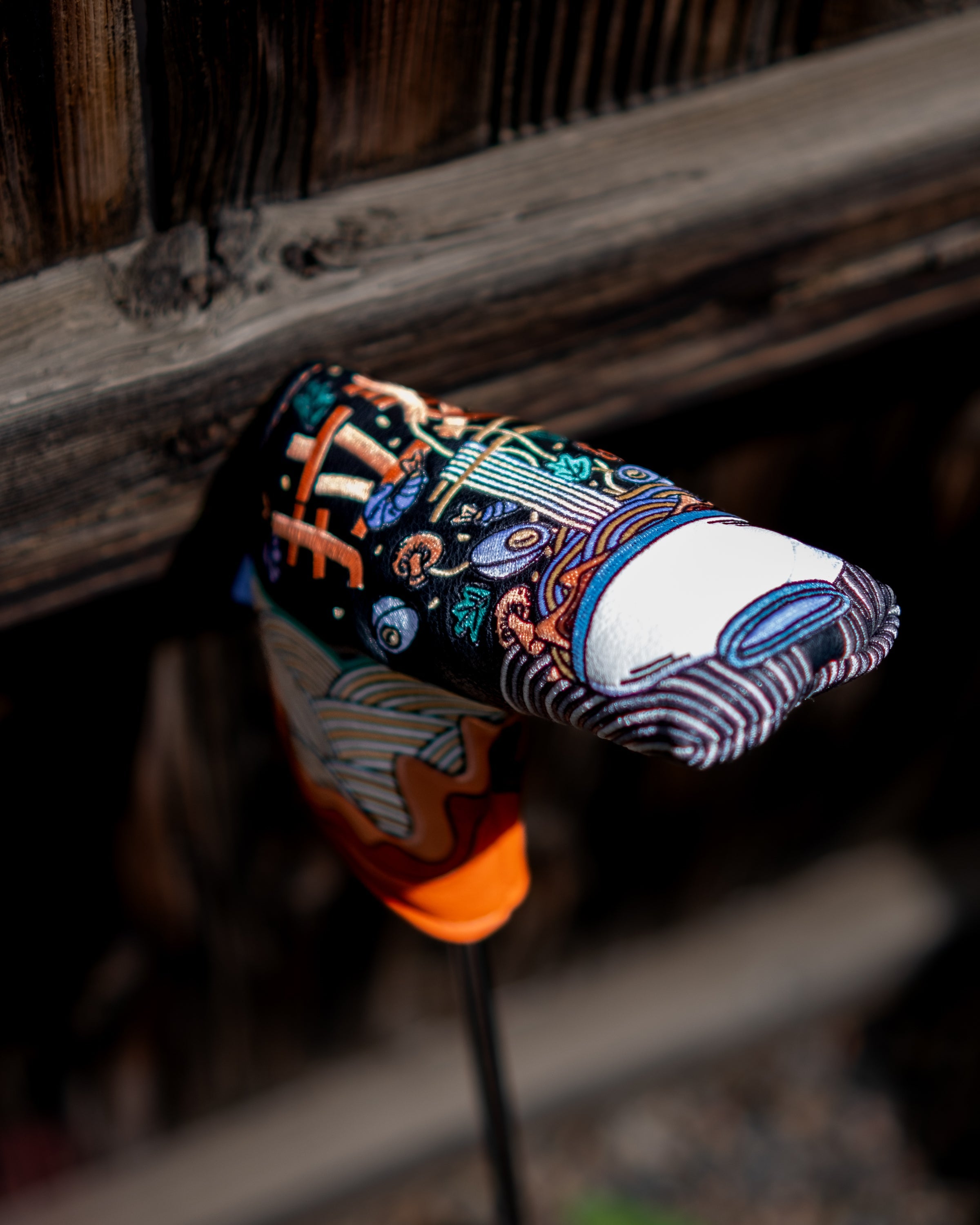 Colorful ramen bowl design Blade Putter Cover resting on a golf club, attached to a golf bag, with a wooden background.