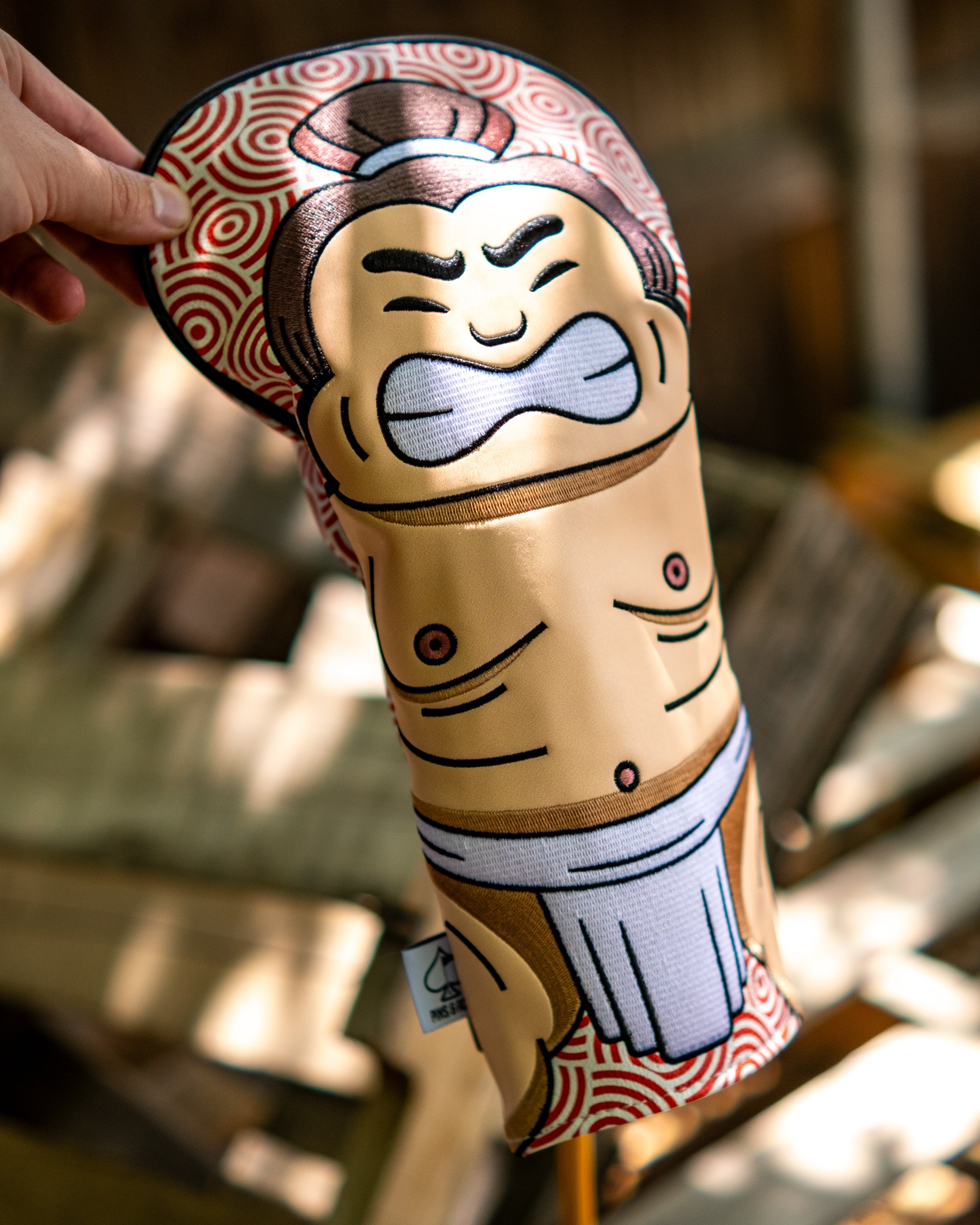 Sumo driver golf club cover featuring a cartoon sumo wrestler with a smiling face, traditional attire, and vibrant red patterned background.