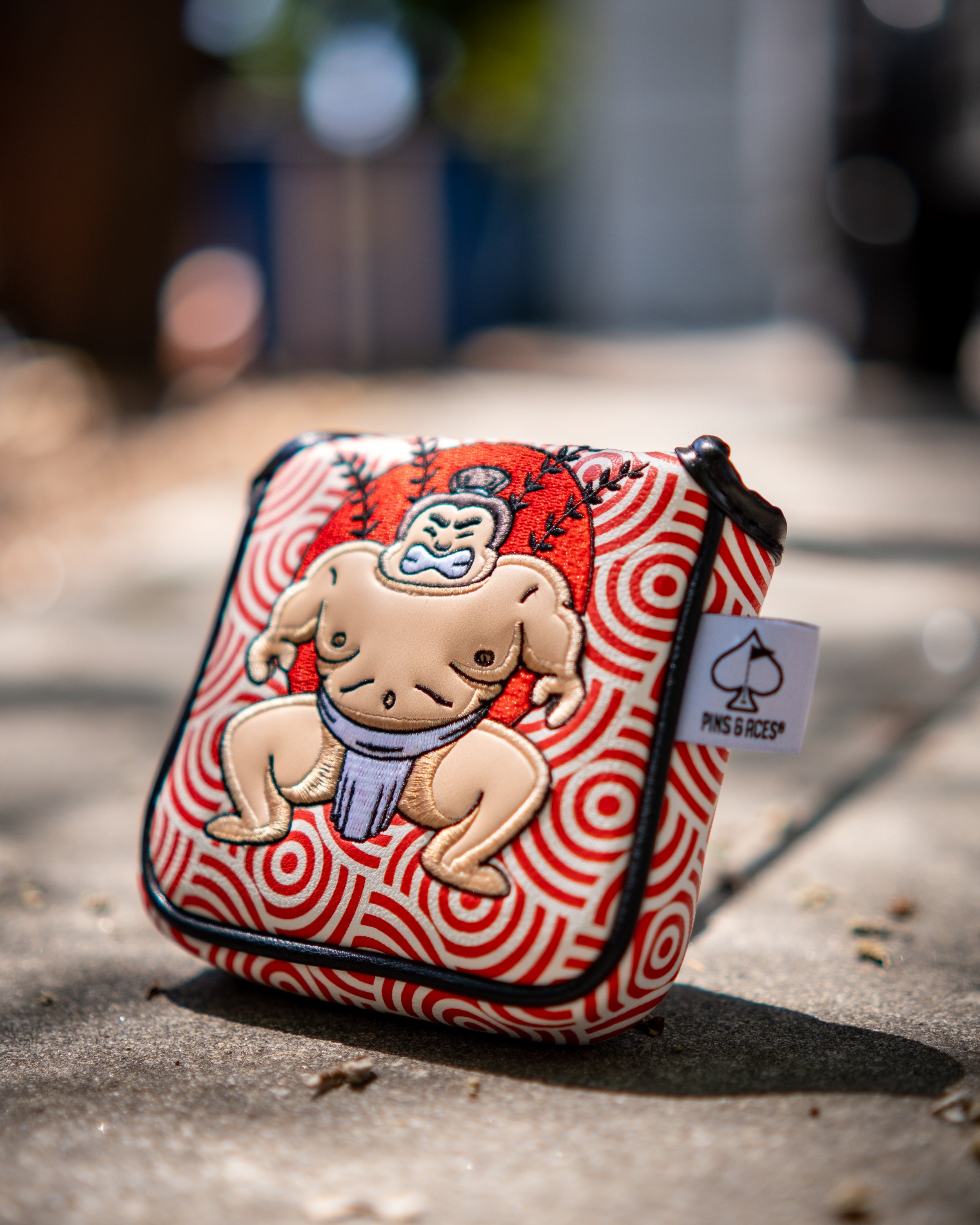 Sumo wrestler embroidered patch on a red and white patterned golf putter cover with a black zipper and a Pins and Aces tag.