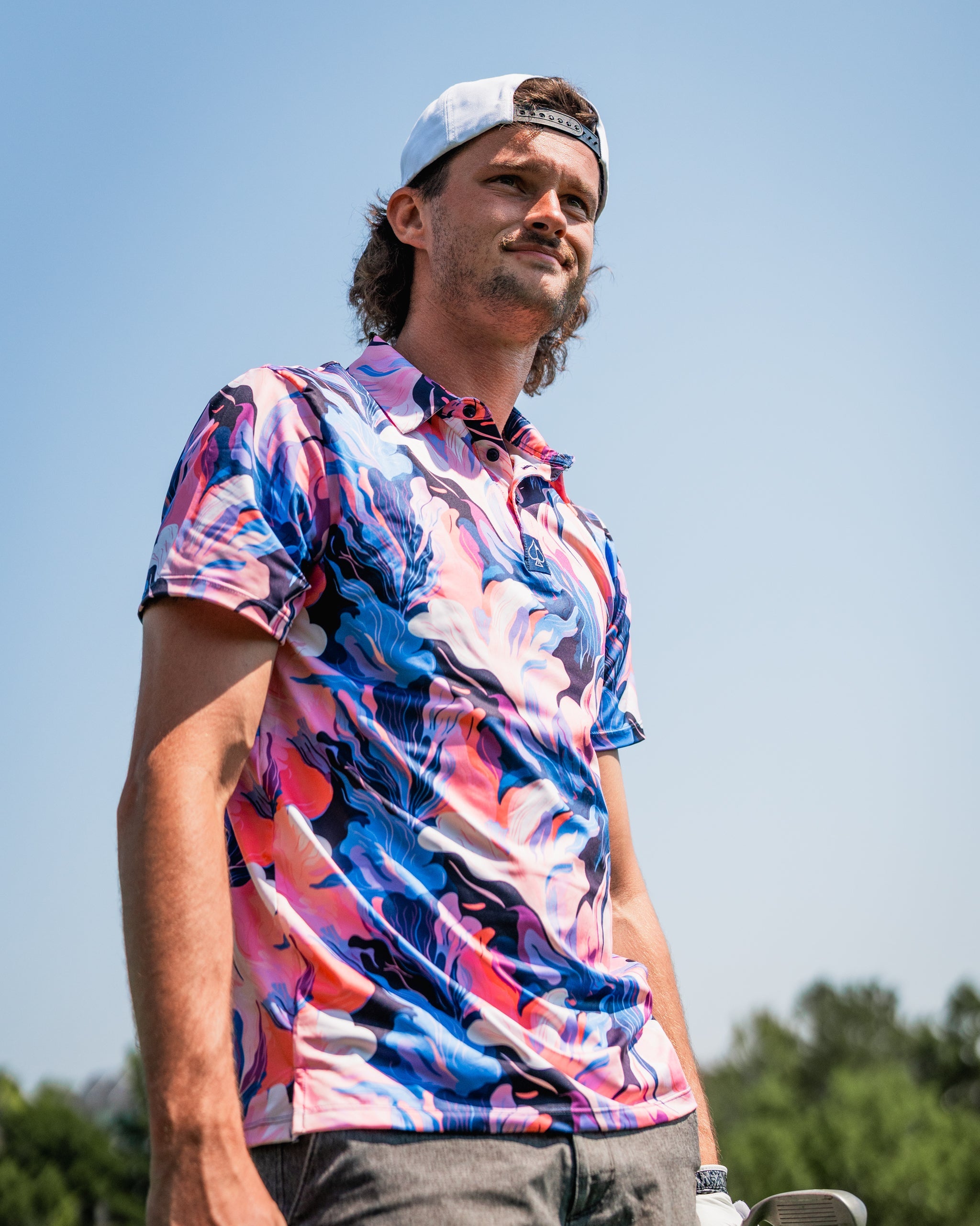 A golfer wearing a vibrant melting floral polo shirt stands confidently on the course under a clear blue sky.
