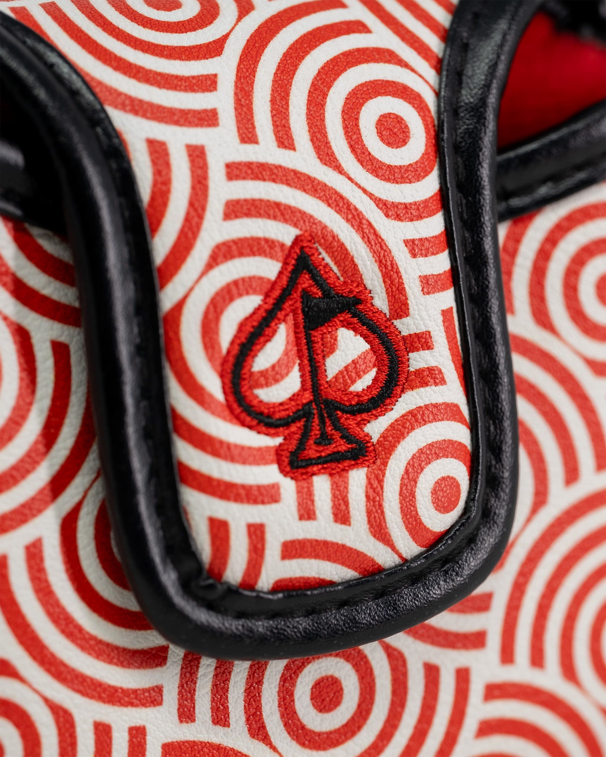Close-up of a golf mallet putter cover featuring a red and white geometric pattern with a black and red spade logo embroidered on it.