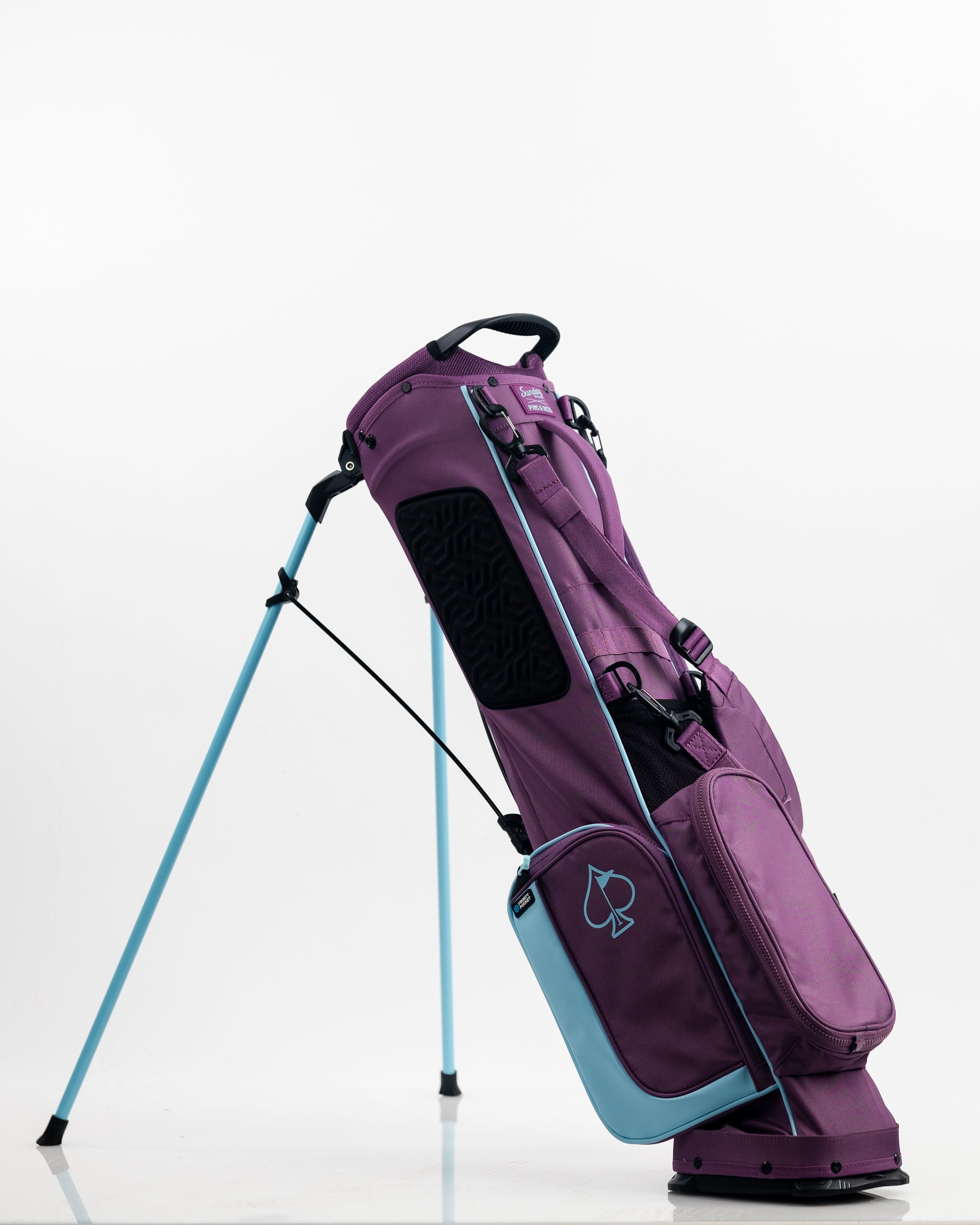 Bold purple golf bag with black, light blue accents, and a spade logo, standing on a blue golf stand against a white backg...