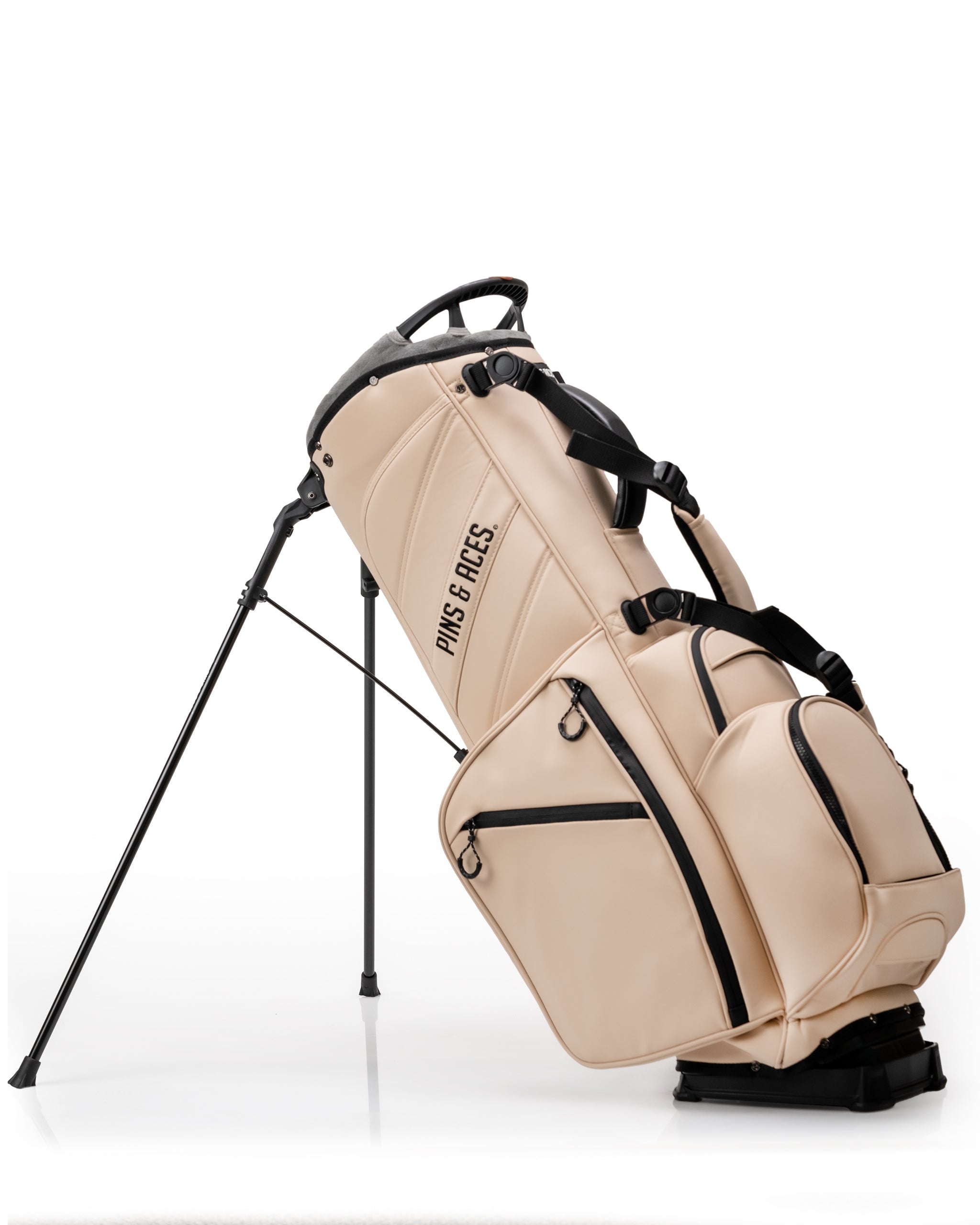 Beige golf bag with black zippers and straps, standing on a black stand, featuring multiple compartments and "Pins & Aces"...