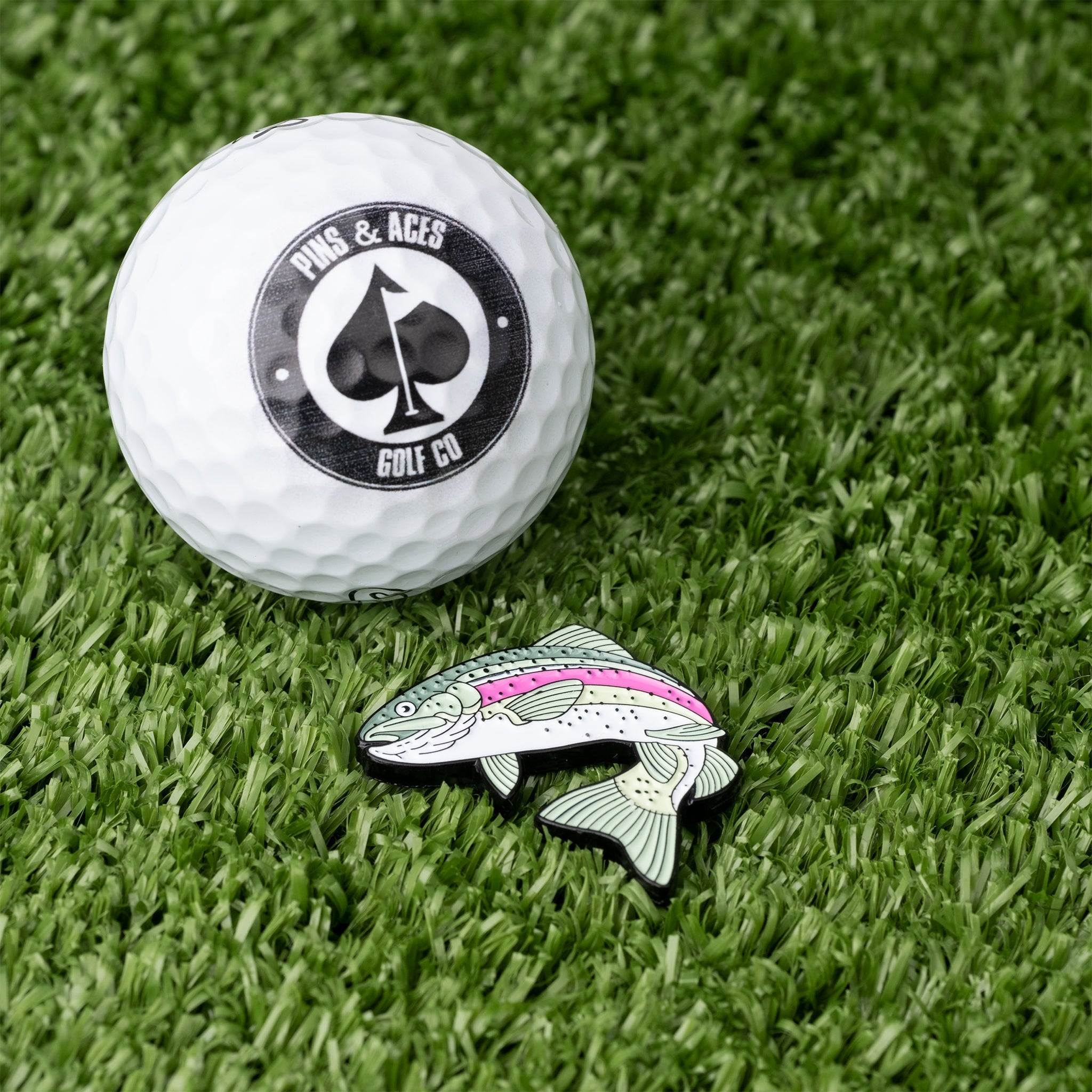 Trout ball marker featuring a detailed trout design, positioned beside a golf ball on a grassy surface.