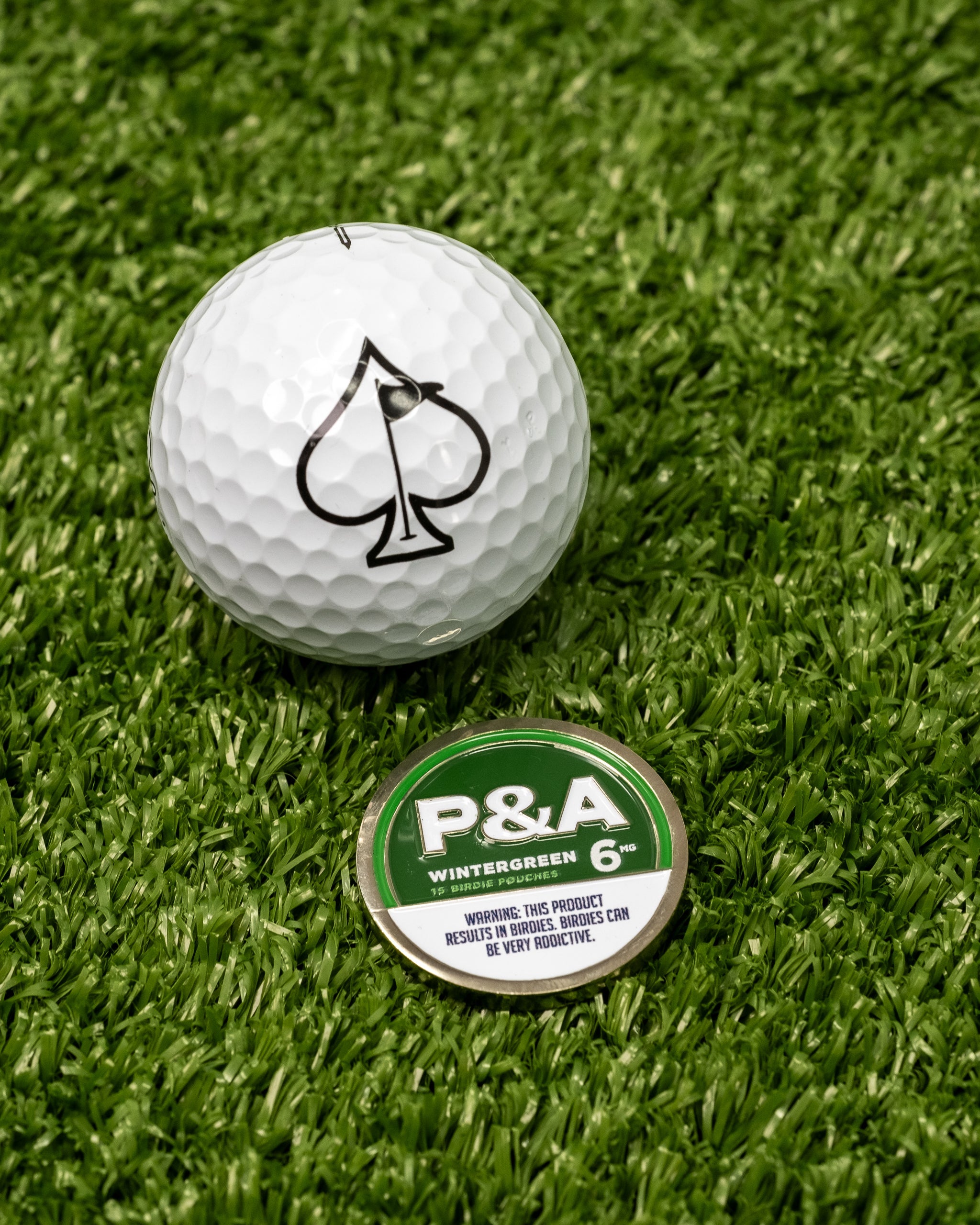 Alt text: "Close-up of a 6 Milly - Wintergreen Ball Marker, featuring a white golf ball with a black spade symbol on artificial grass next to a green and silver Wintergreen 6 marker disc with warning text."