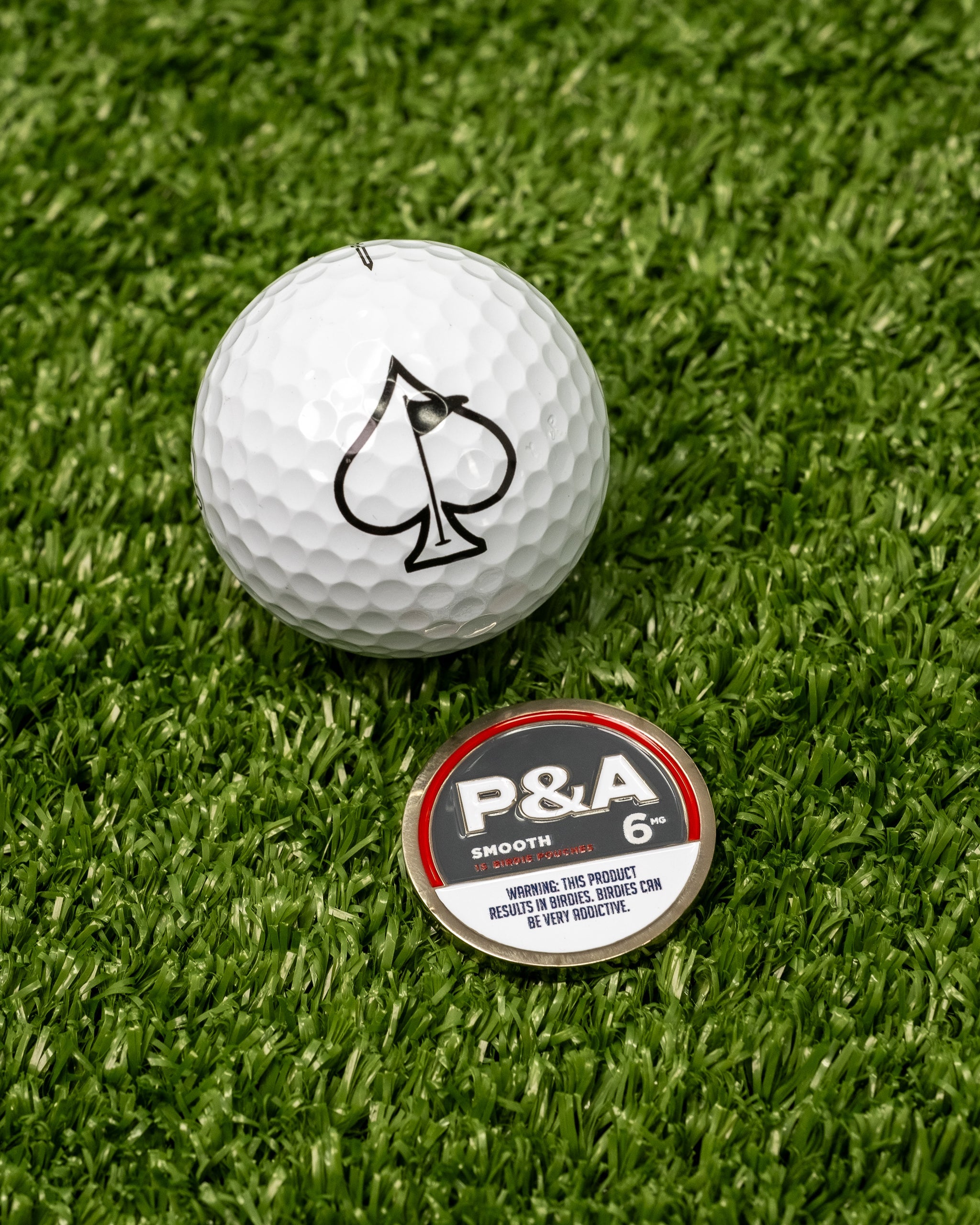 White golf ball with black spade logo next to a round marker labeled "P&A 6" on green turf.