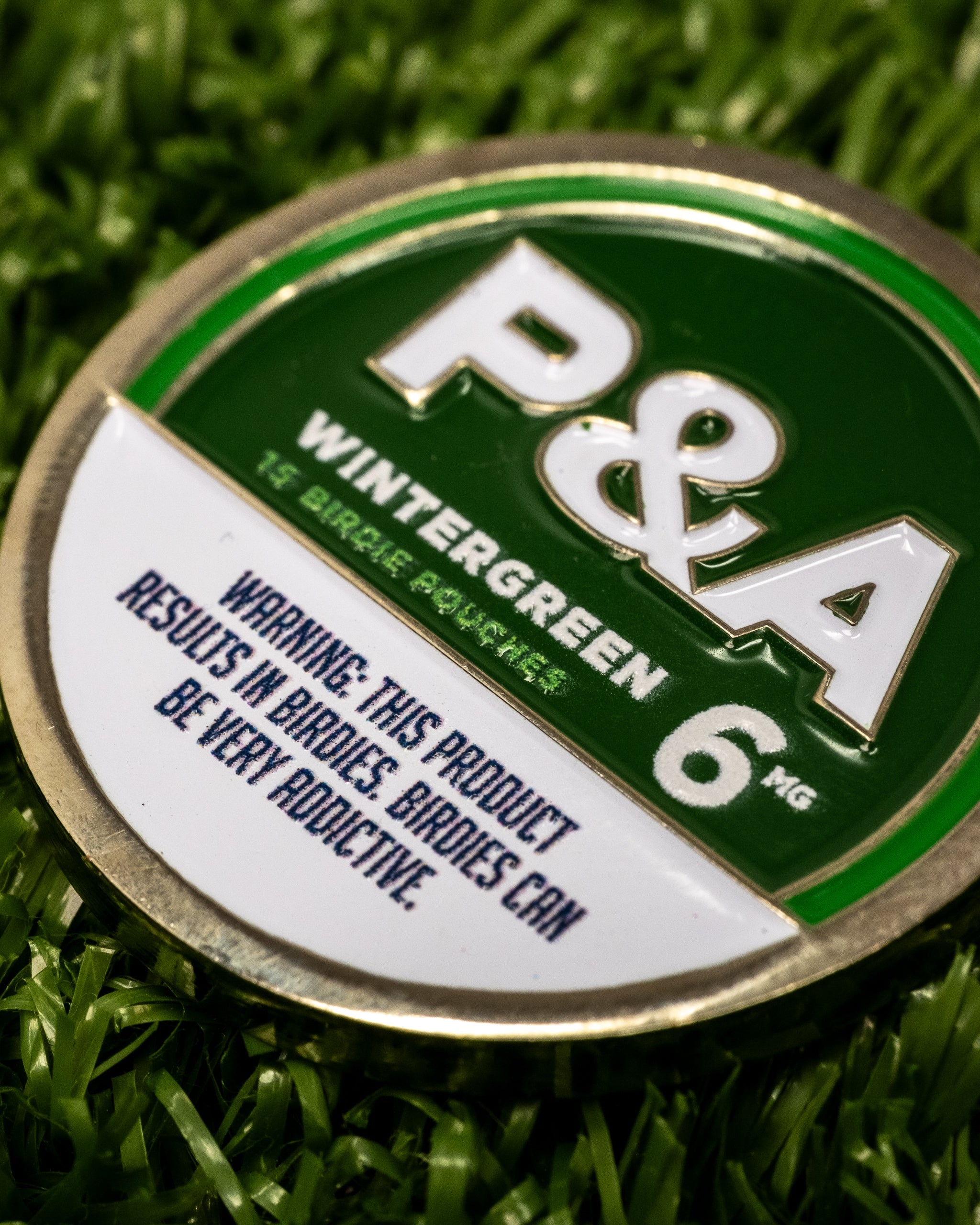 Close-up of a 6-milligram Milly Wintergreen golf ball marker placed on artificial grass, featuring a bold green and white color scheme with the text "P&A Wintergreen," and a warning label indicating the product is addictive.