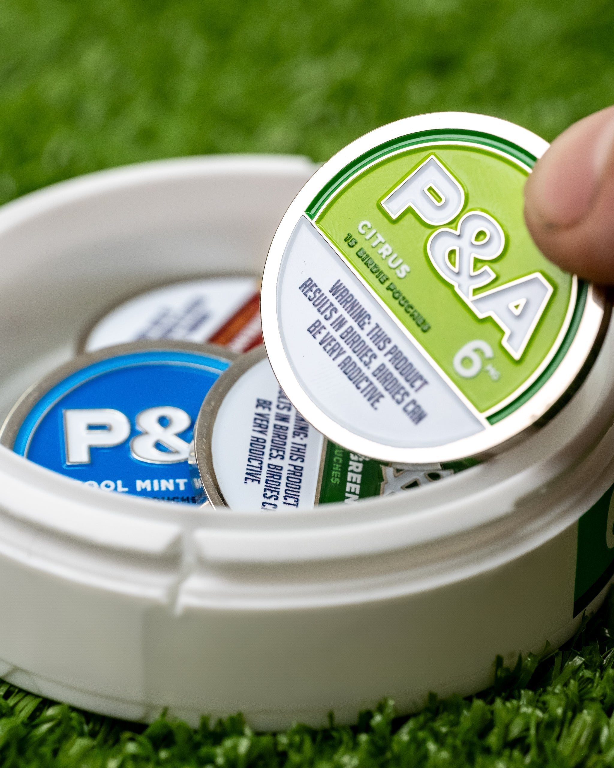 Hand holding a 6 Milly Citrus ball marker from a container with various colored markers on green turf.