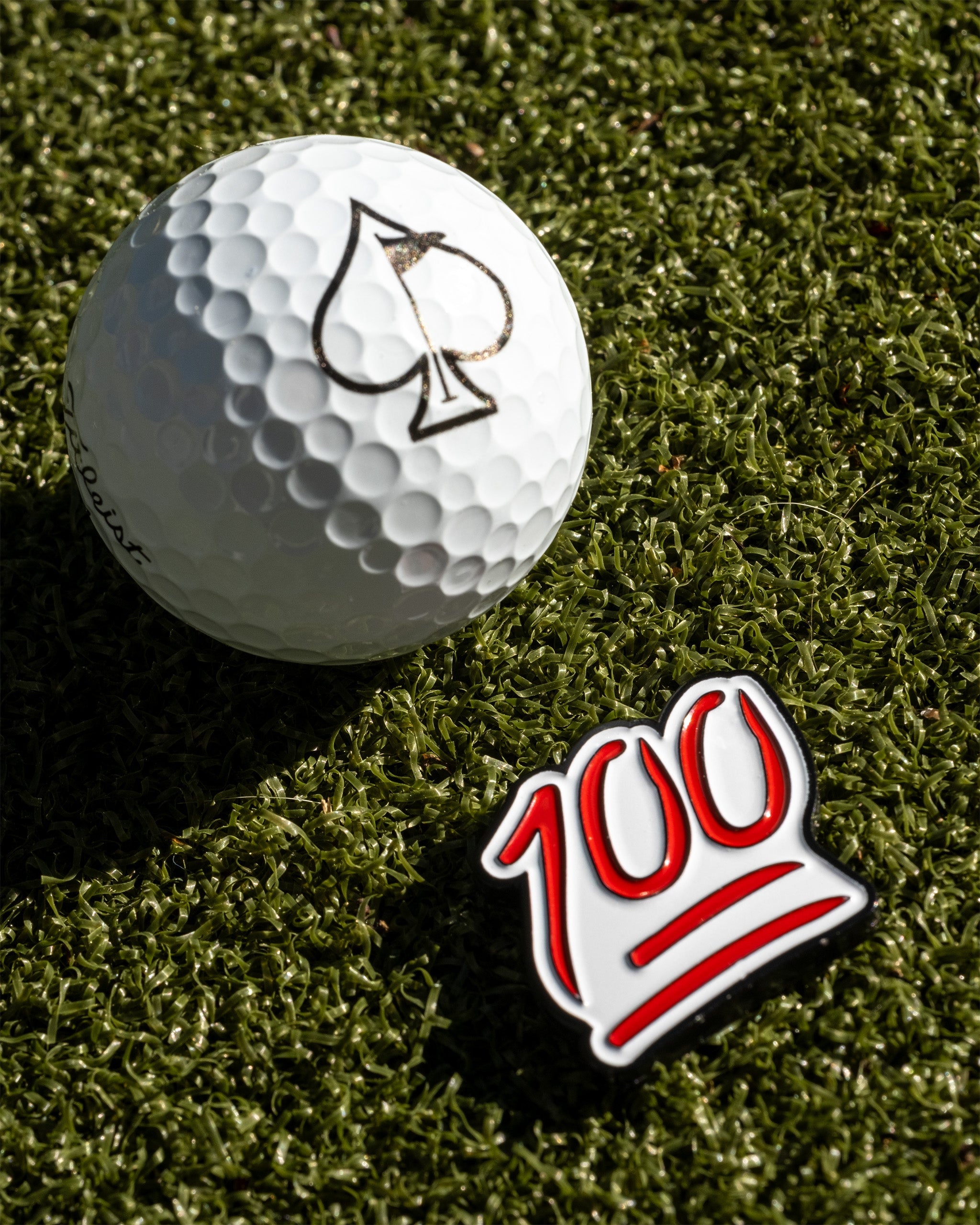 Bold golf ball marker featuring a vibrant "100" design, placed next to a golf ball with a spade logo on a green turf background.
