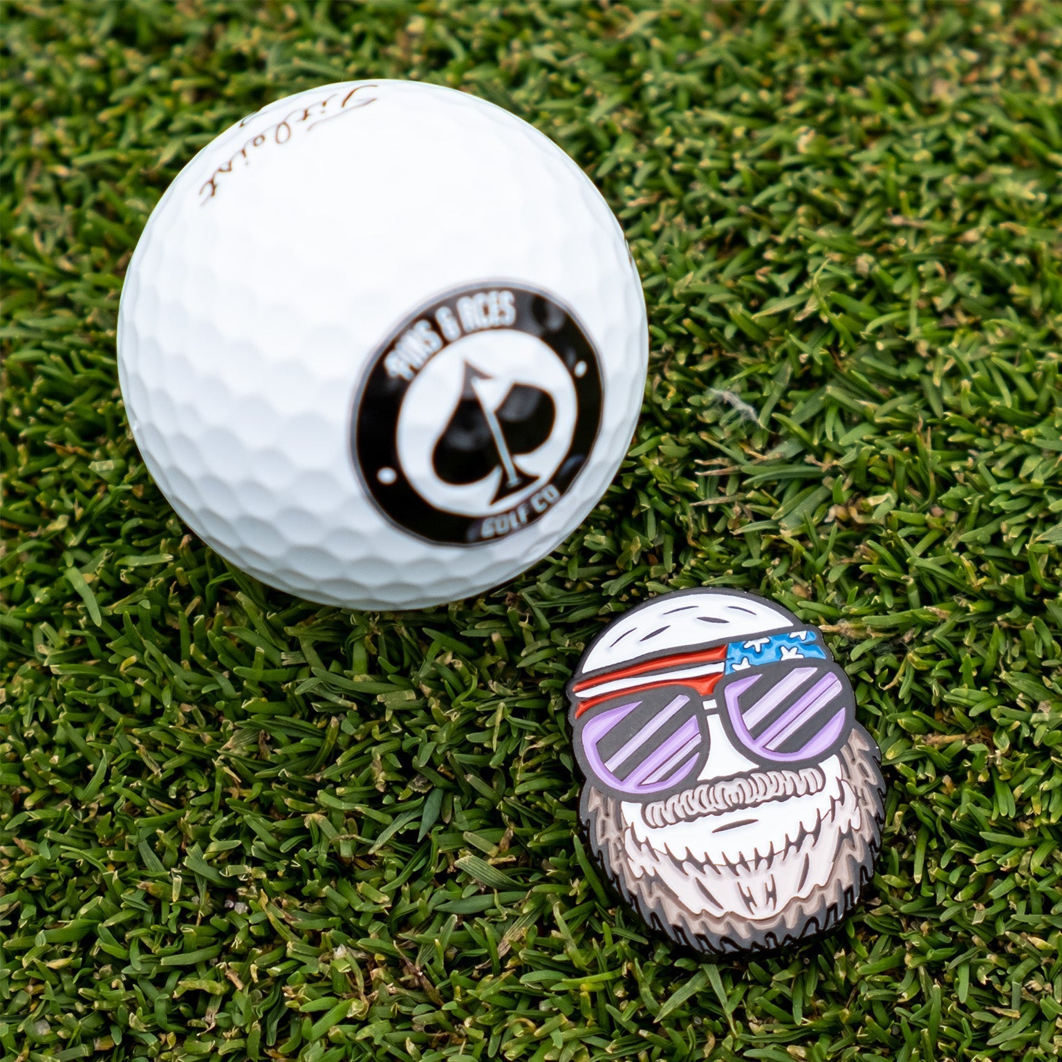 Gus the Degenerate Ball Marker features a playful design with sunglasses and a beard, placed next to a golf ball on grass.
