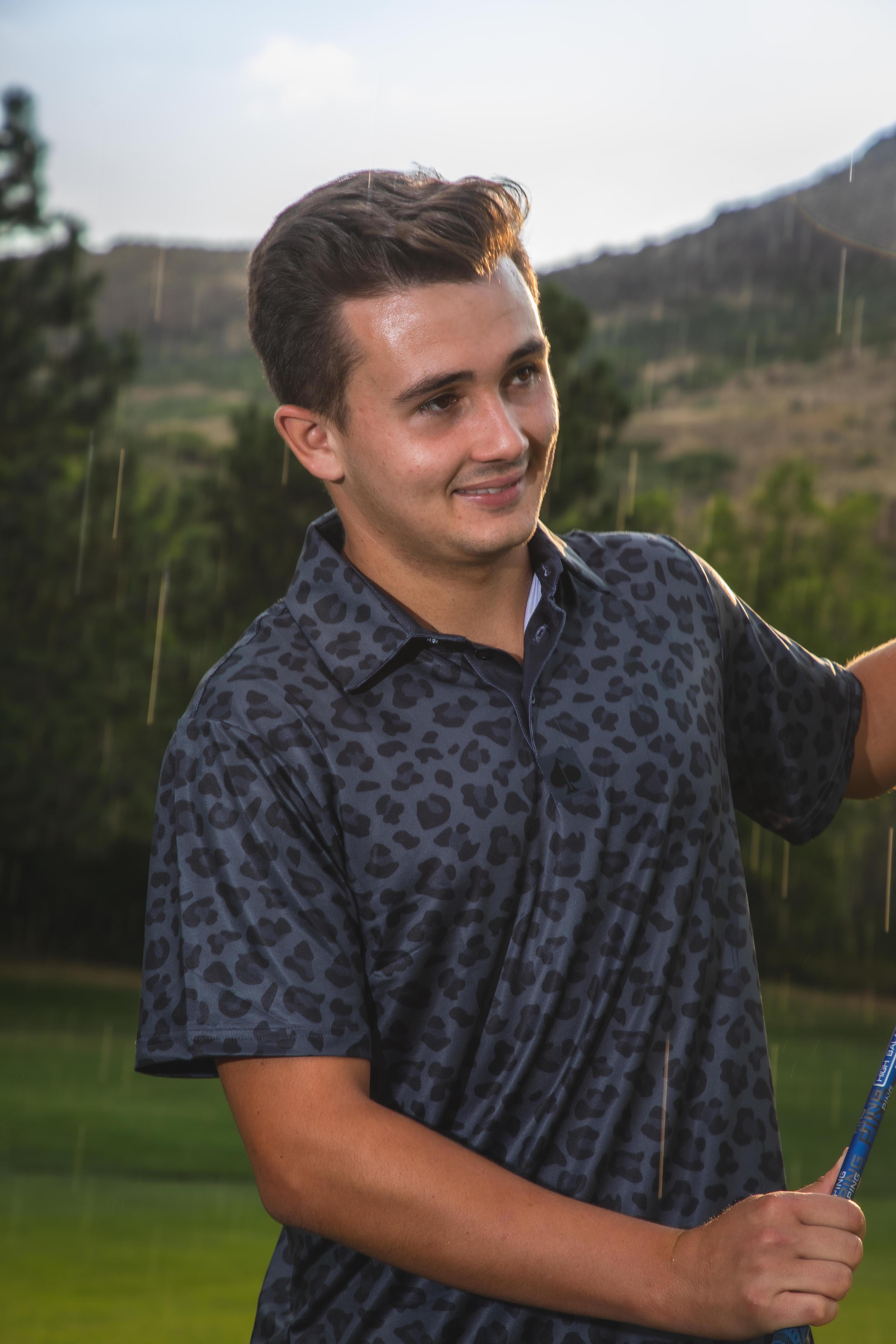 A golfer wearing a black jungle cat print polo shirt smiles while holding a golf club on a rainy day outdoors.