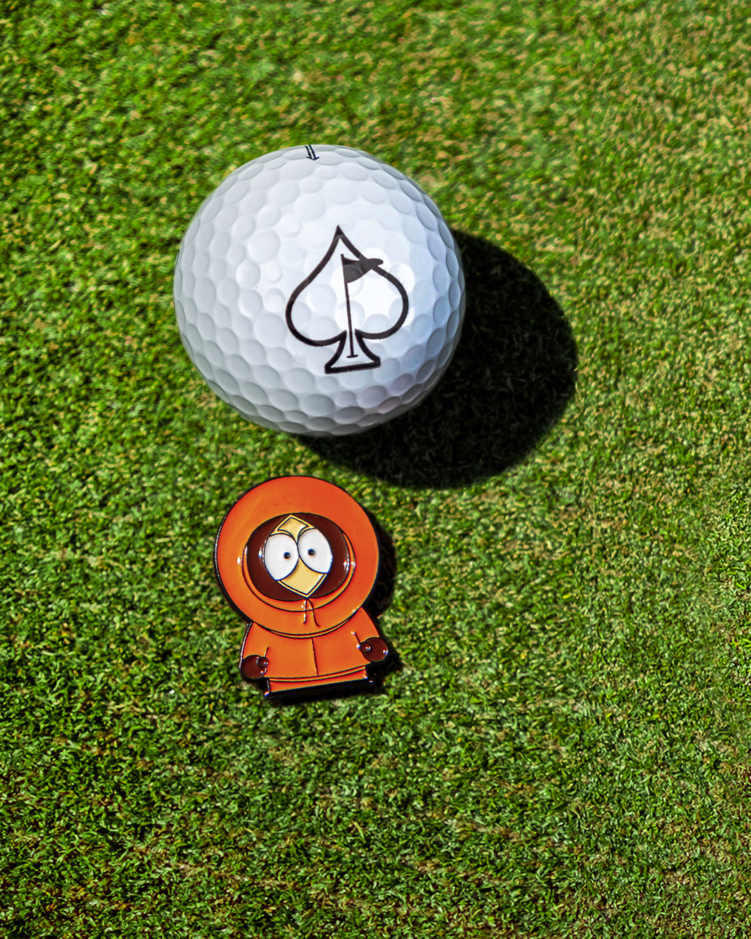 Kenny from South Park ball marker next to a golf ball with an ace of spades logo on a green golf course.