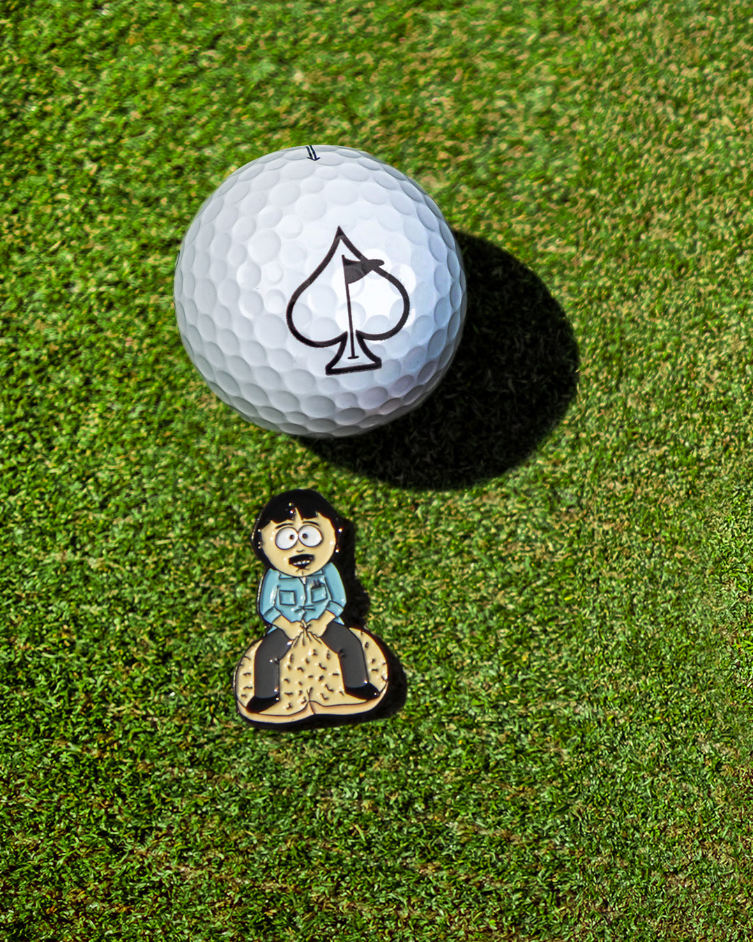South Park Randy ball marker beside a golf ball on green grass, perfect for adding personality to your game.