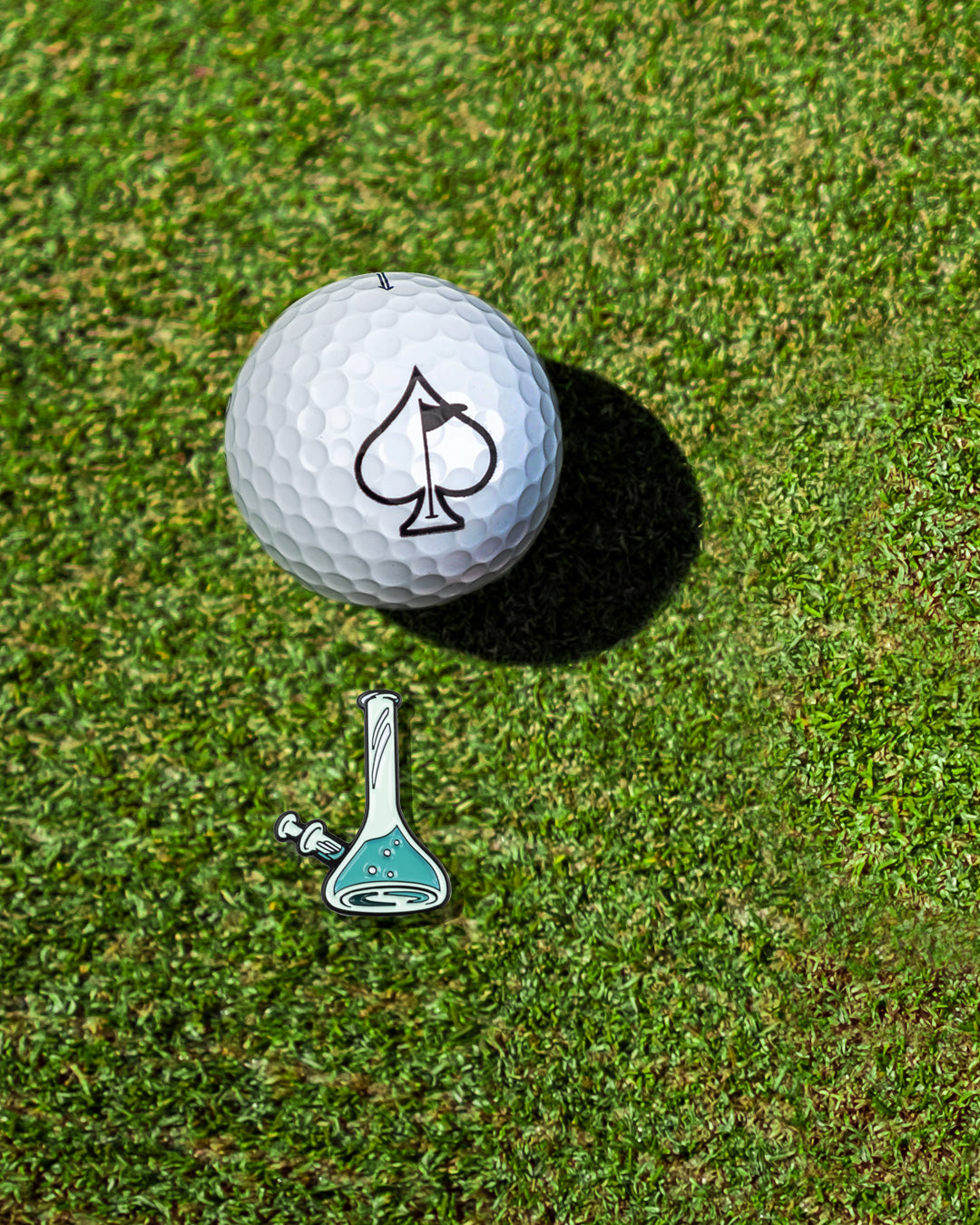 A golf ball with a spade logo on a grass surface next to a bong-shaped ball marker.