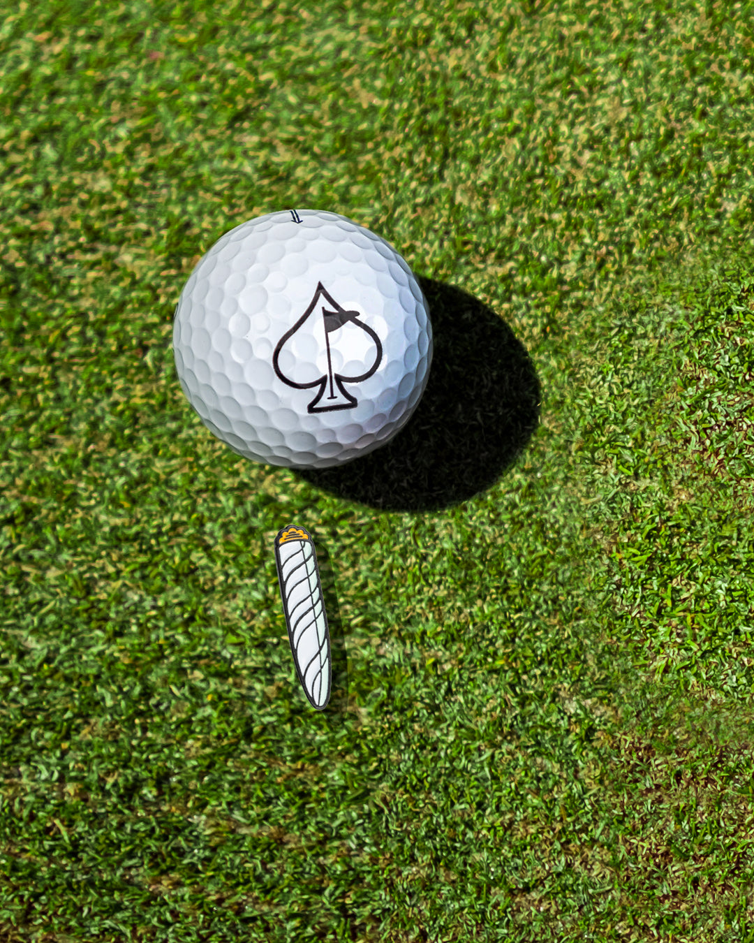 Alt text: "Close-up view of a white golf ball with a black spade design on top next to a novelty 420-themed joint-shaped ball marker on a grassy background."