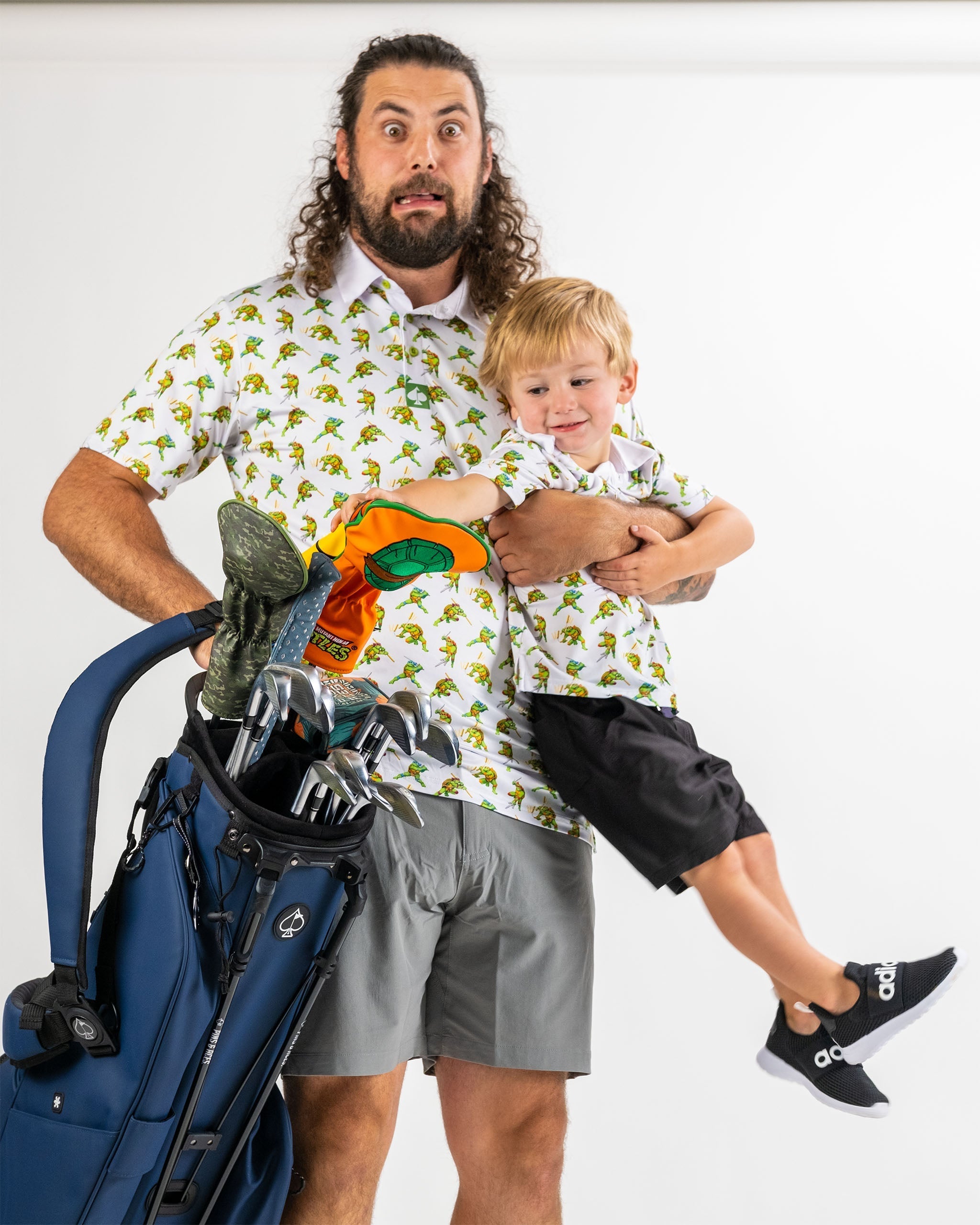 Father and son wearing matching TMNT Cowabunga Classic 8-Bit shirts, holding golf clubs and a bag, showcasing playful style.