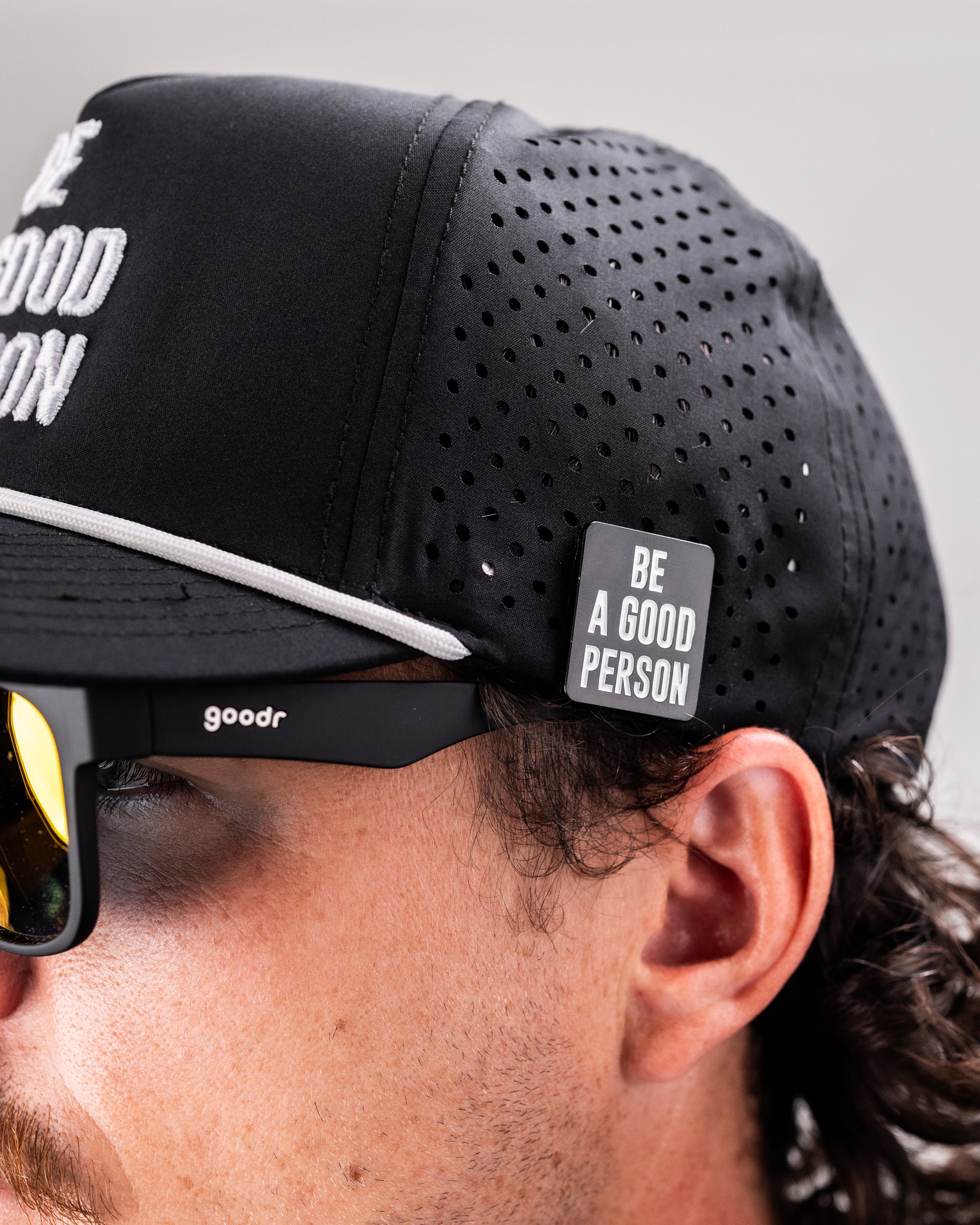 Black cap with "Be A Good Person" text, featuring a ball marker attached to the side, worn by a person with sunglasses.
