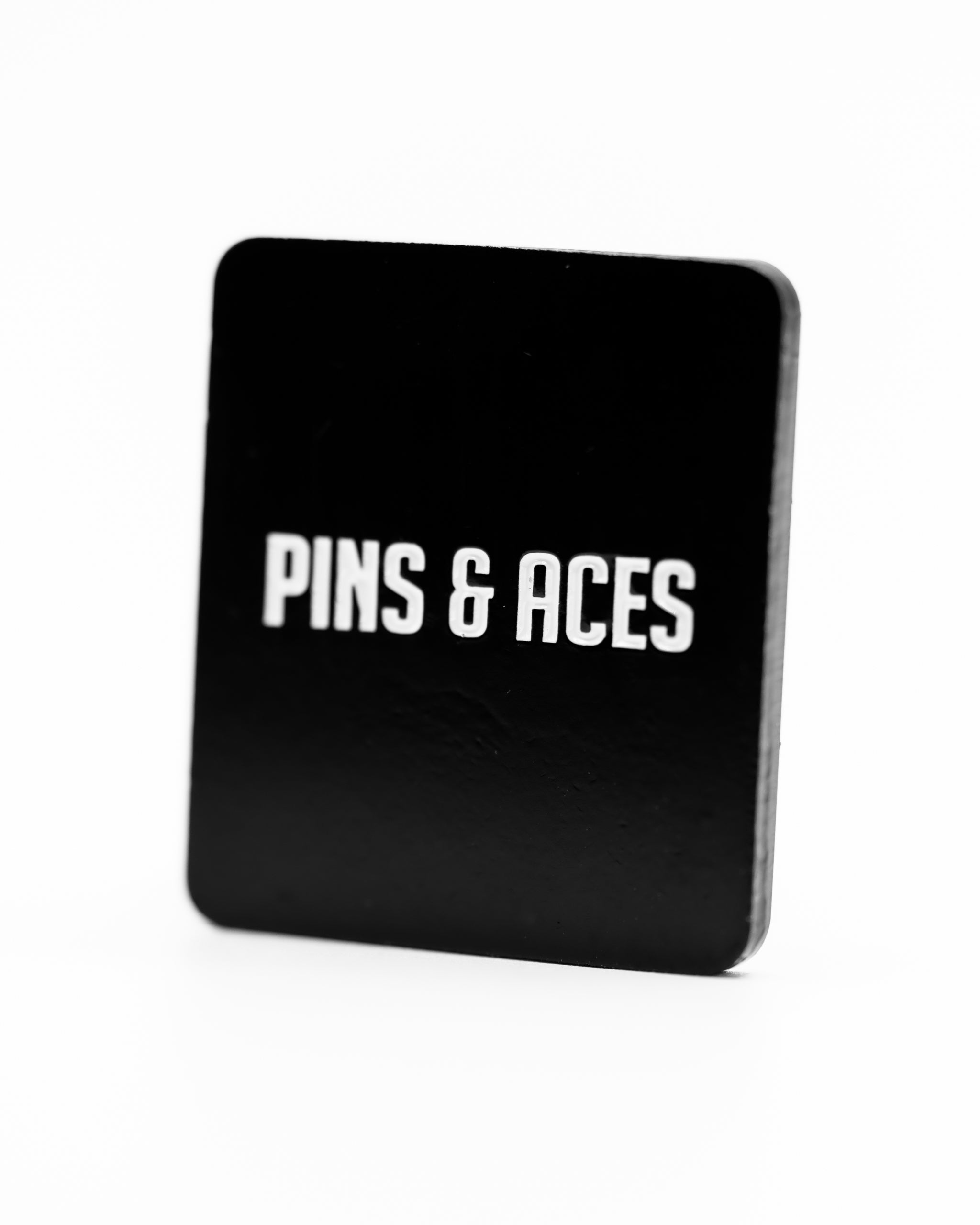 Black ball marker featuring the text "Pins & Aces" in bold white letters, designed for golfers who value positivity.