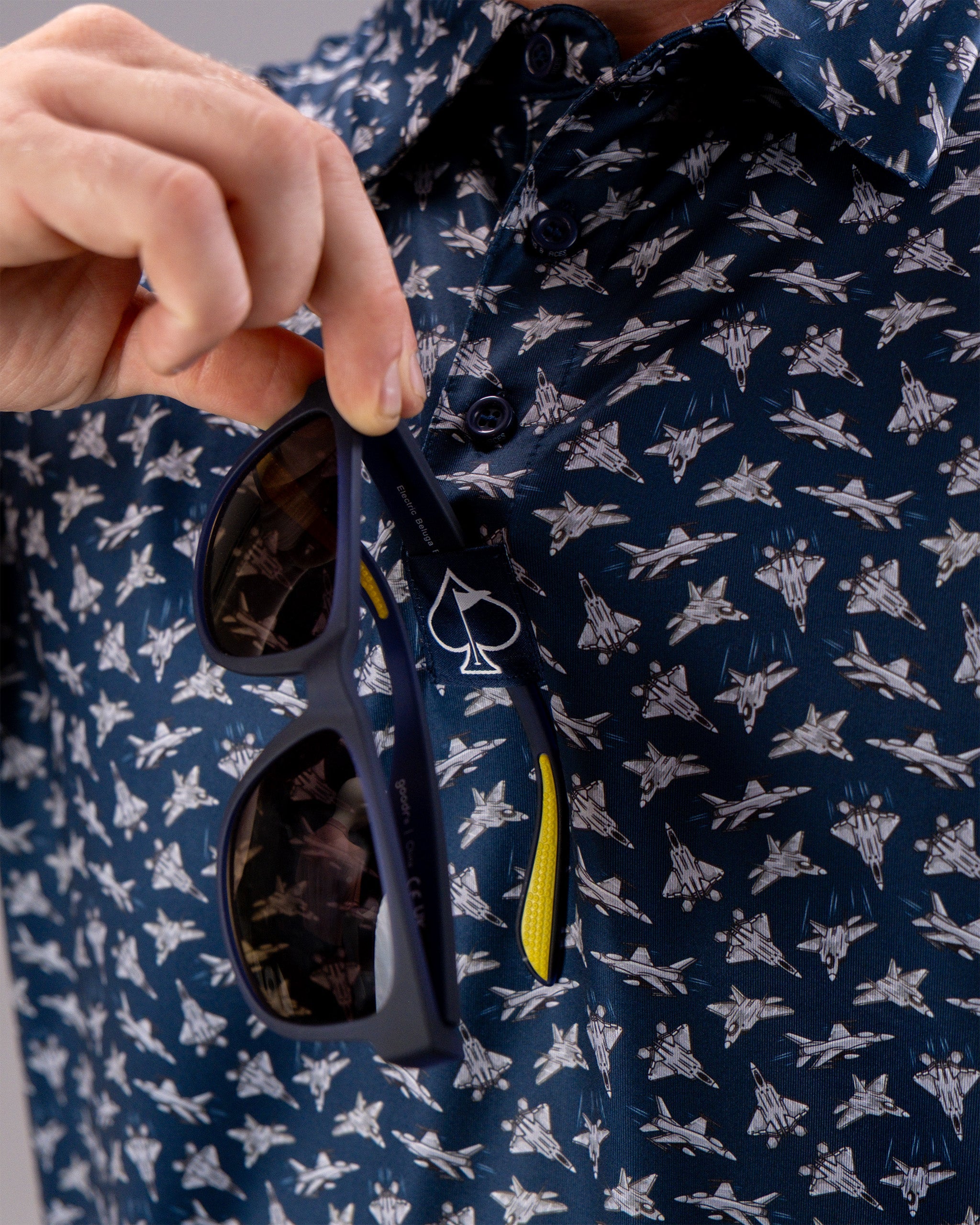 Close-up of a person holding sunglasses with a Birds of Steel logo tag, wearing a navy shirt with a jet aircraft pattern.