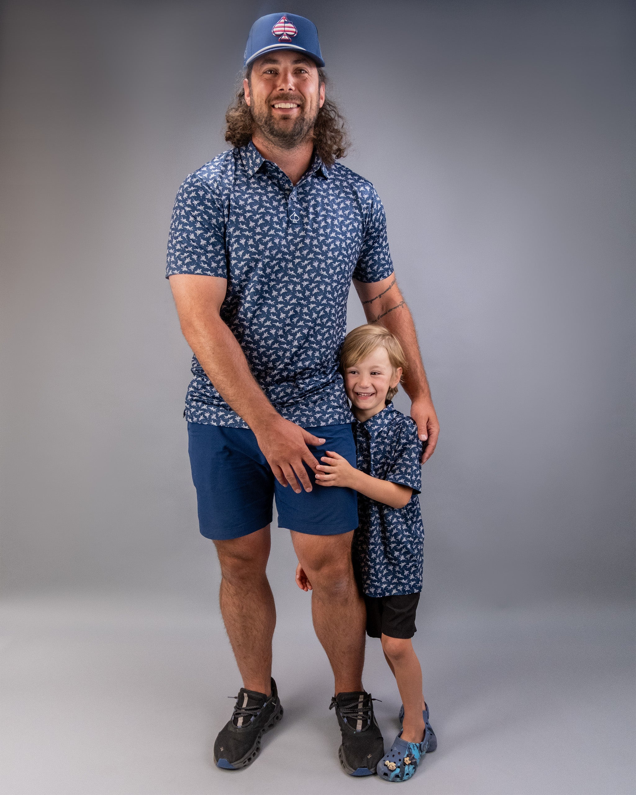 A man and a young boy smiling and hugging, both wearing matching blue patterned shirts, standing against a neutral gray background.