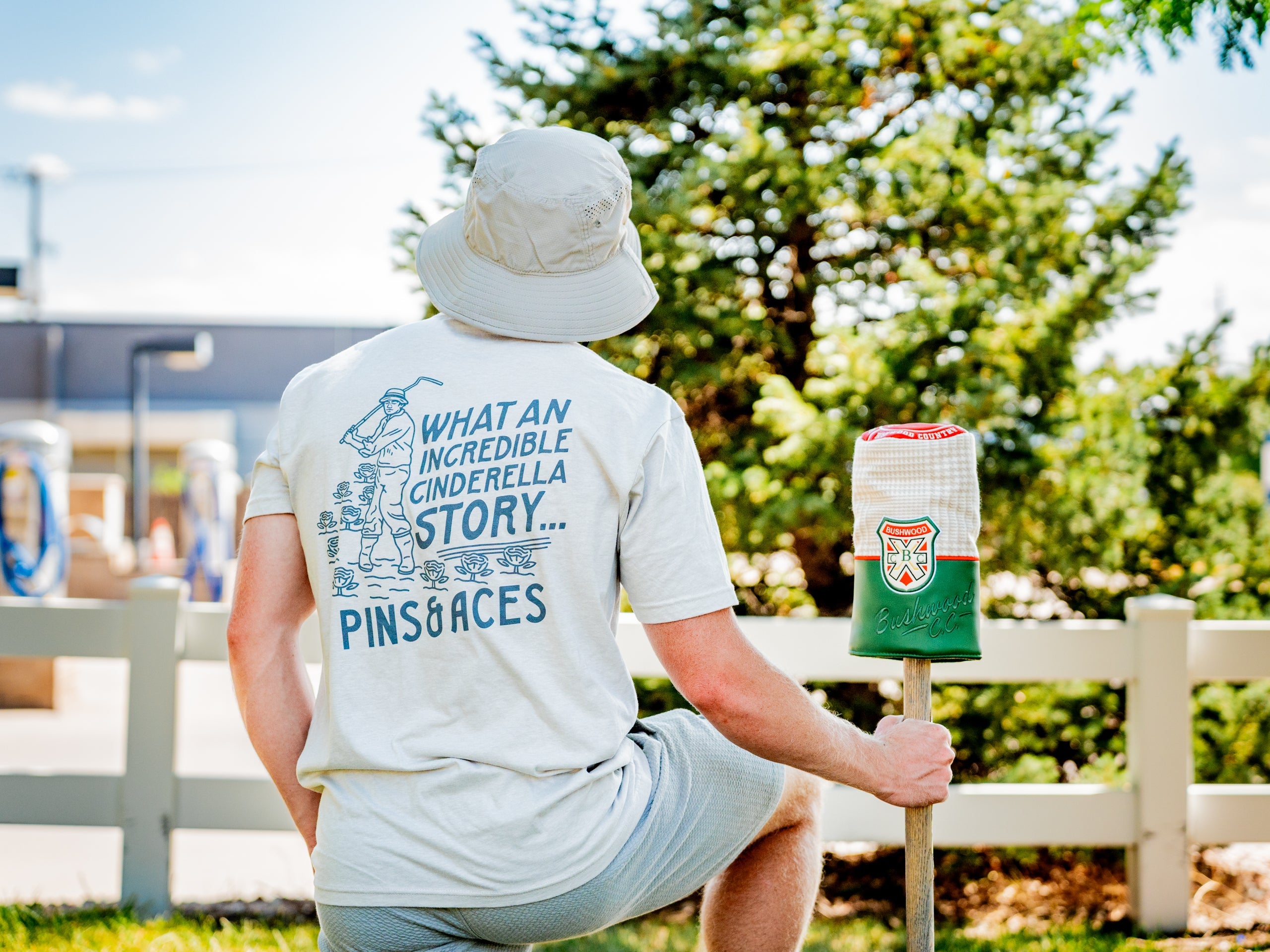 Man wearing a light gray Bushwood Crest 3.0 tee shirt with a whimsical golf design, sitting beside a golf headcover.