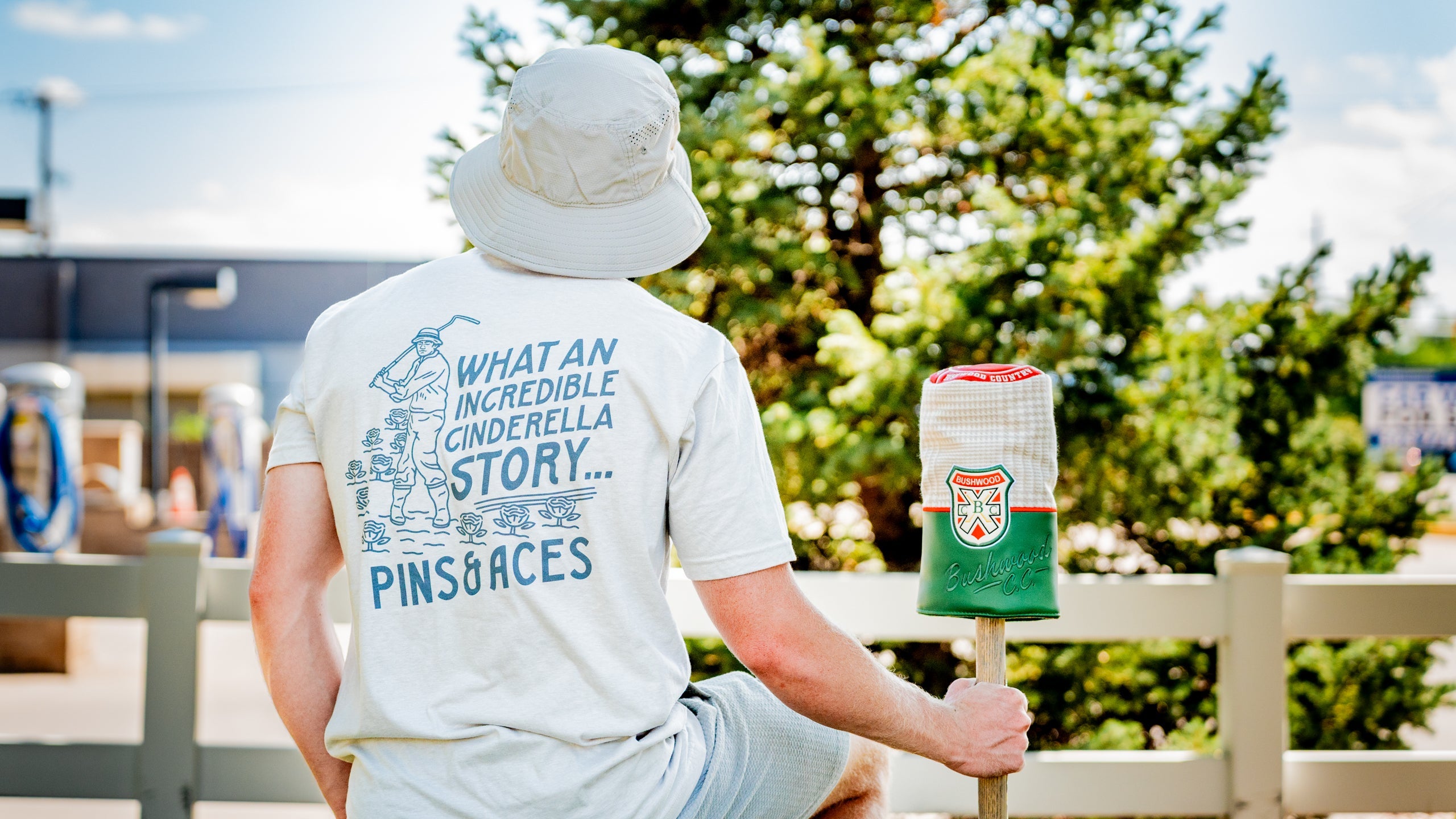 Man sitting on a fence wearing a light gray Bushwood Crest 3.0 tee shirt featuring a whimsical golf graphic on the back.
