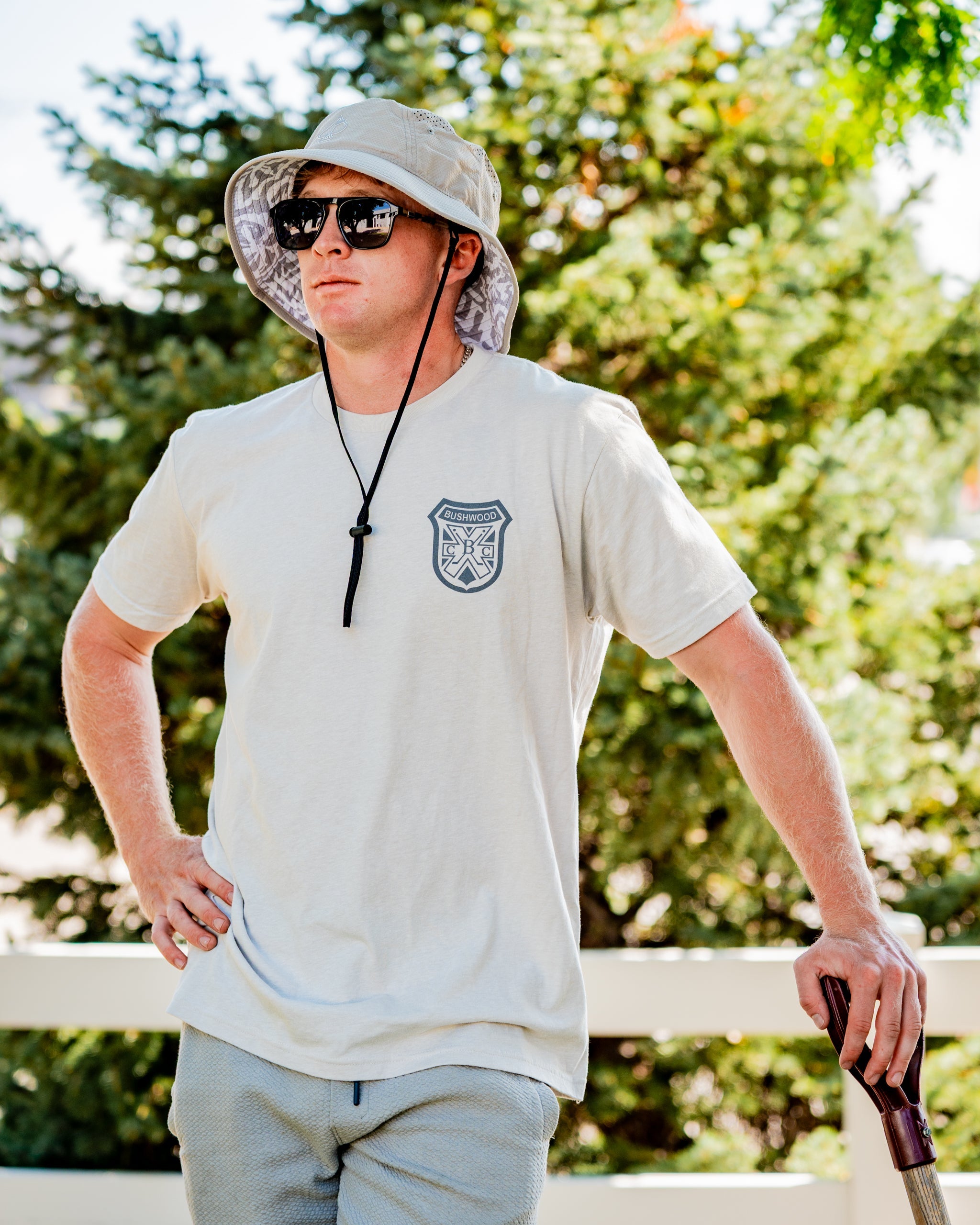 Man wearing a light-colored Bushwood Crest 3.0 tee shirt, sunglasses, and a bucket hat, posing confidently outdoors.