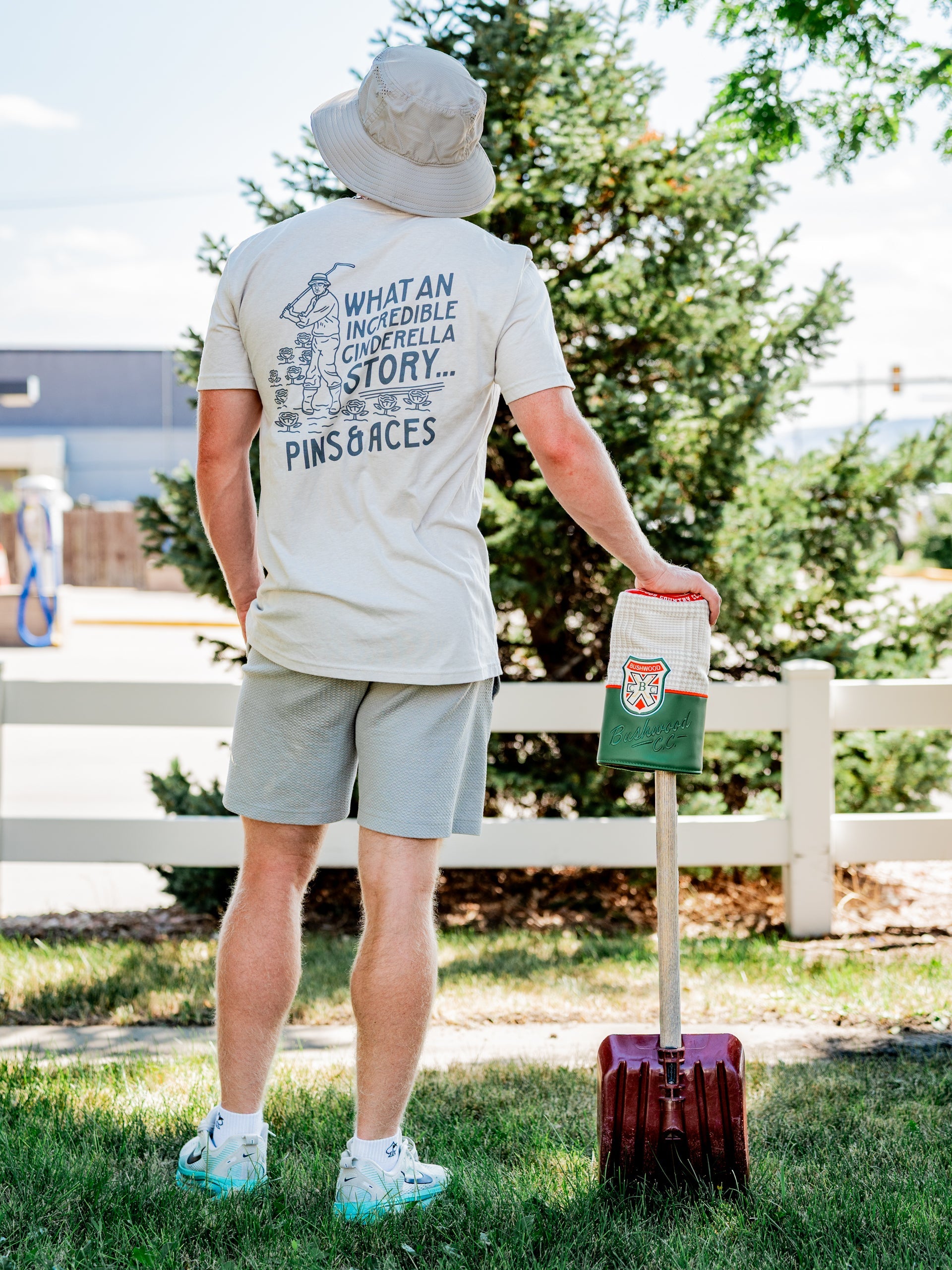 Man wearing a light-colored Bushwood Crest 3.0 tee shirt with a whimsical graphic on the back, standing by a golf bag.