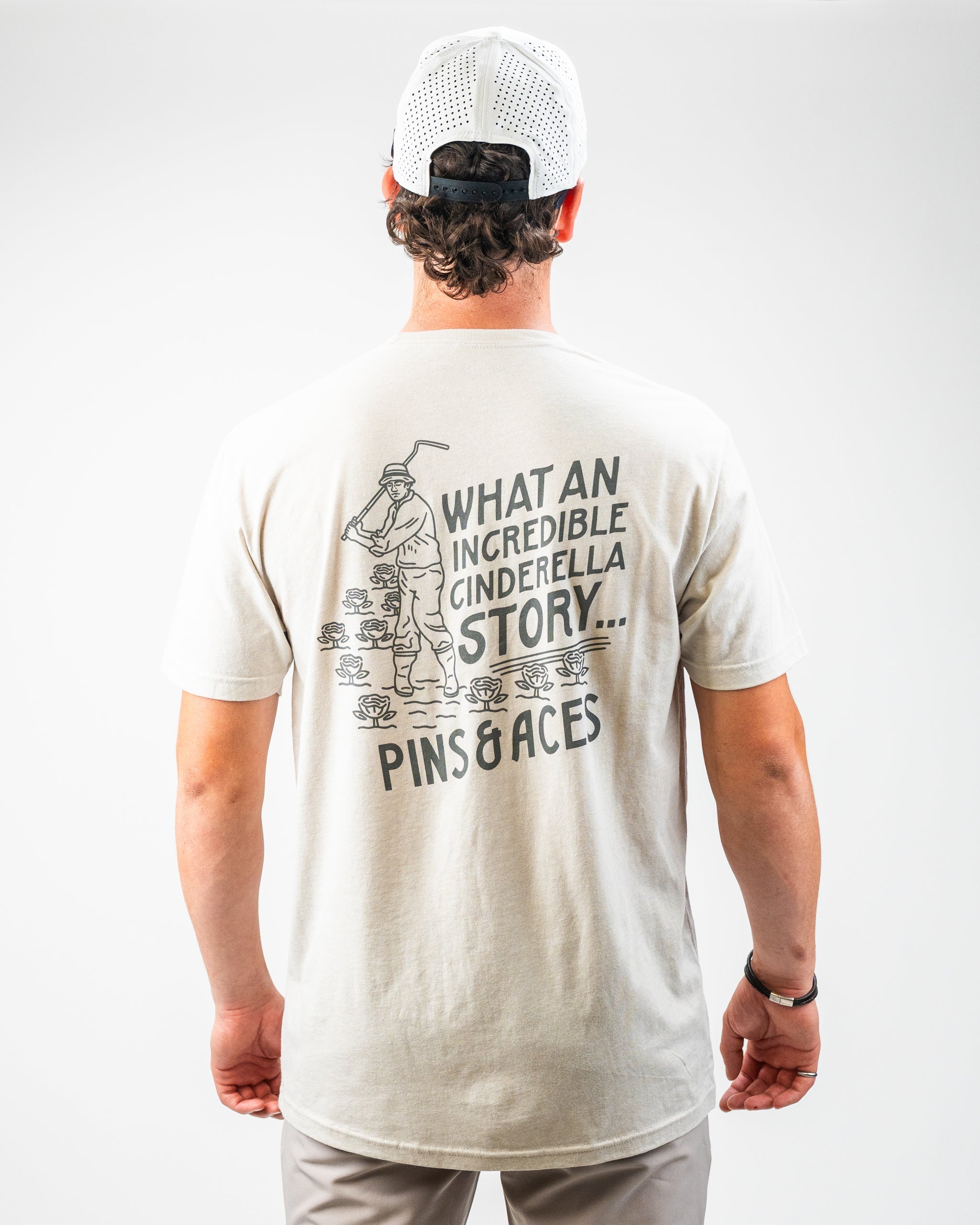 Back view of a cream-colored Bushwood Crest 3.0 tee shirt featuring a golfer and the text "What an Incredible Cinderella Story" by Pins and Aces.