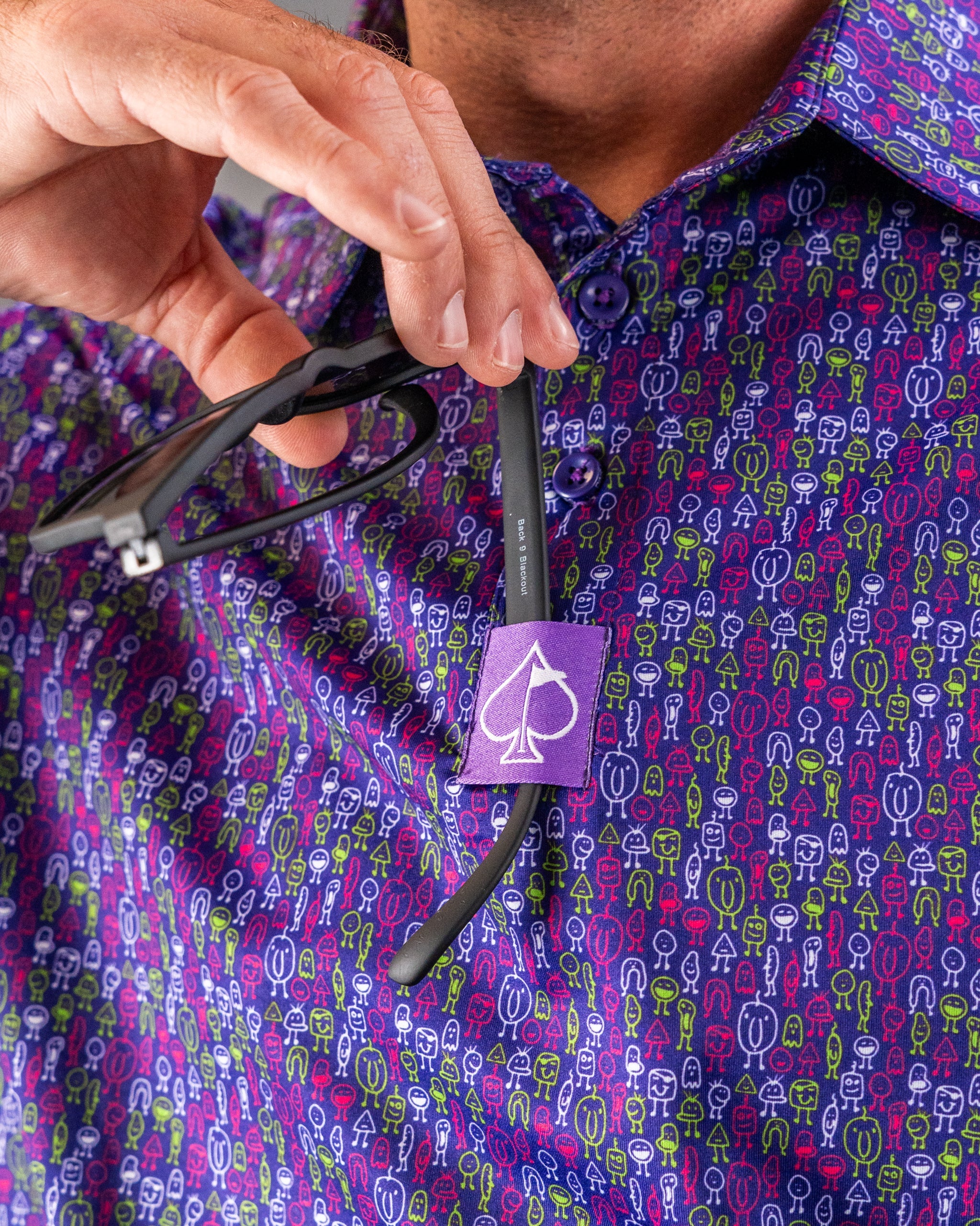 Close-up of a person holding black glasses on a colorful, patterned golf shirt with a purple spade logo tag, highlighting ...