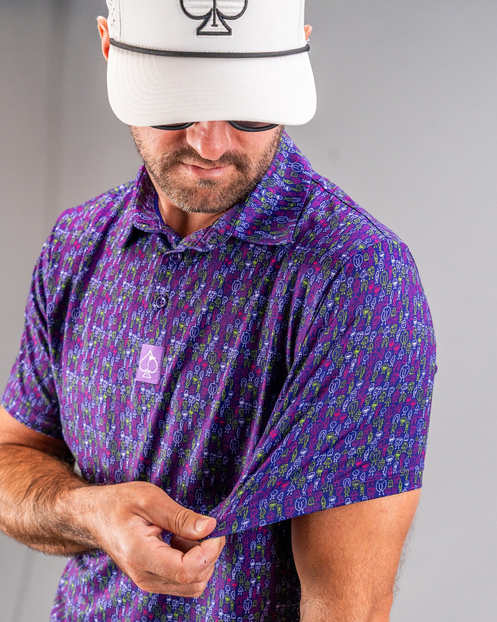 Man wearing a colorful purple golf shirt with quirky, bold patterns and a white cap with a black spade logo.