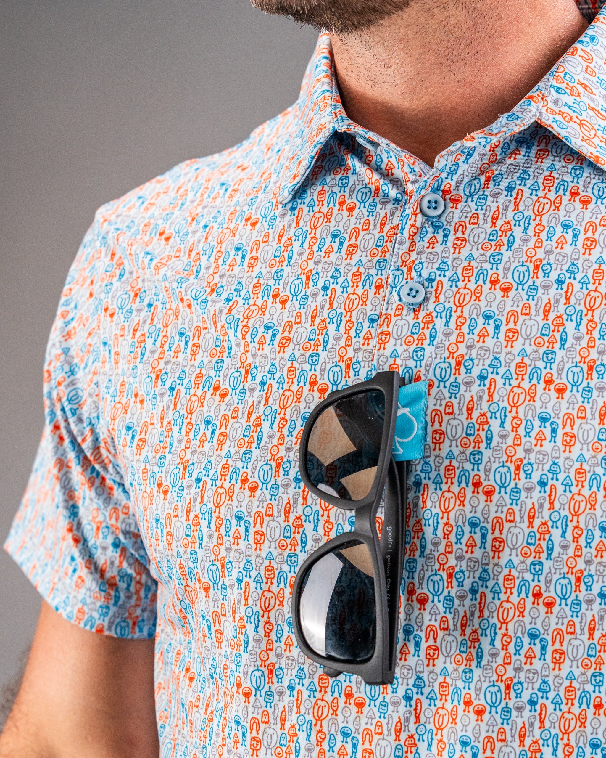 Colorful patterned golf shirt with blue, orange, and gray cartoon characters, paired with sunglasses clipped on the chest.