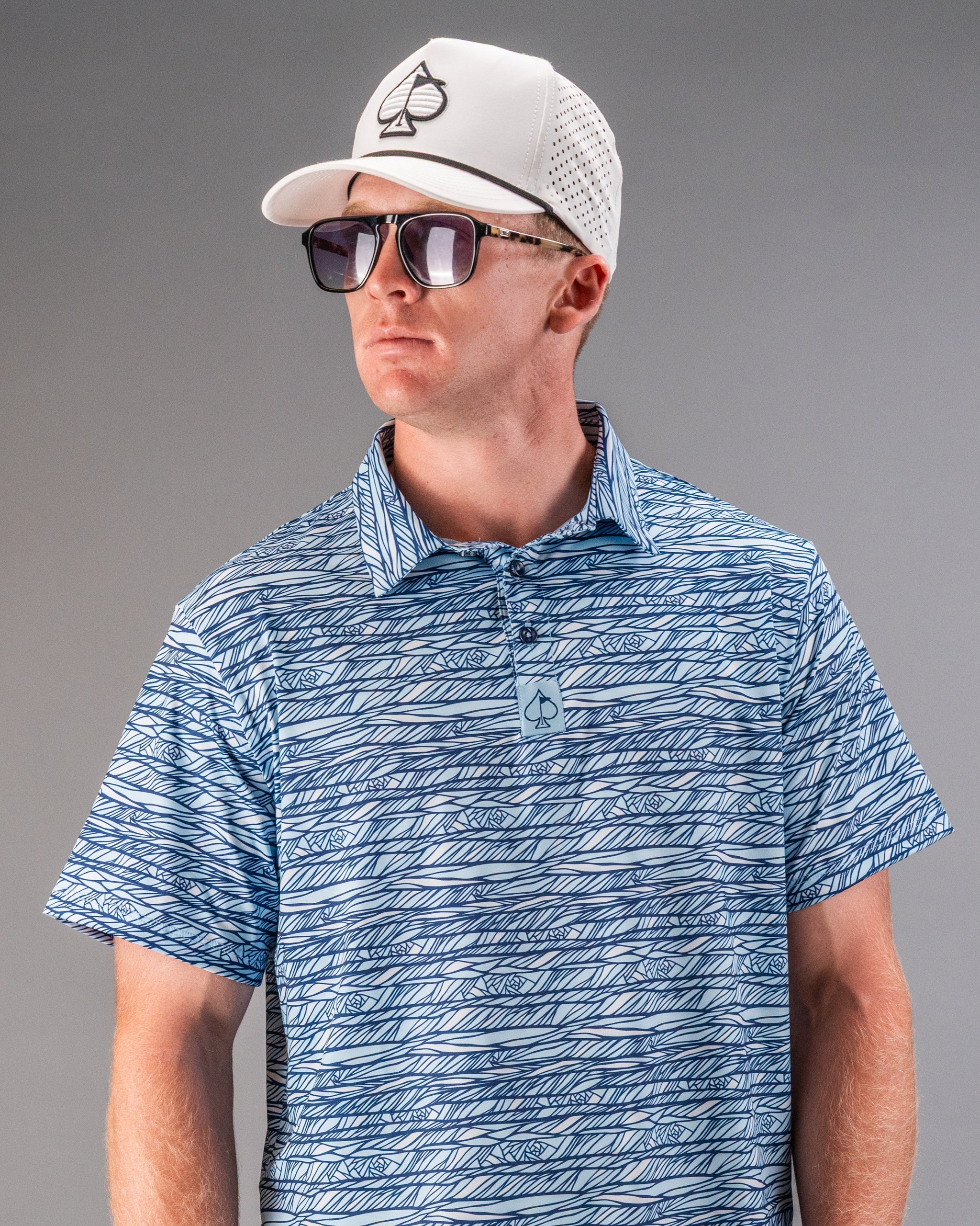 Male model in a blue and white patterned golf shirt, white cap with spade logo, and sunglasses.