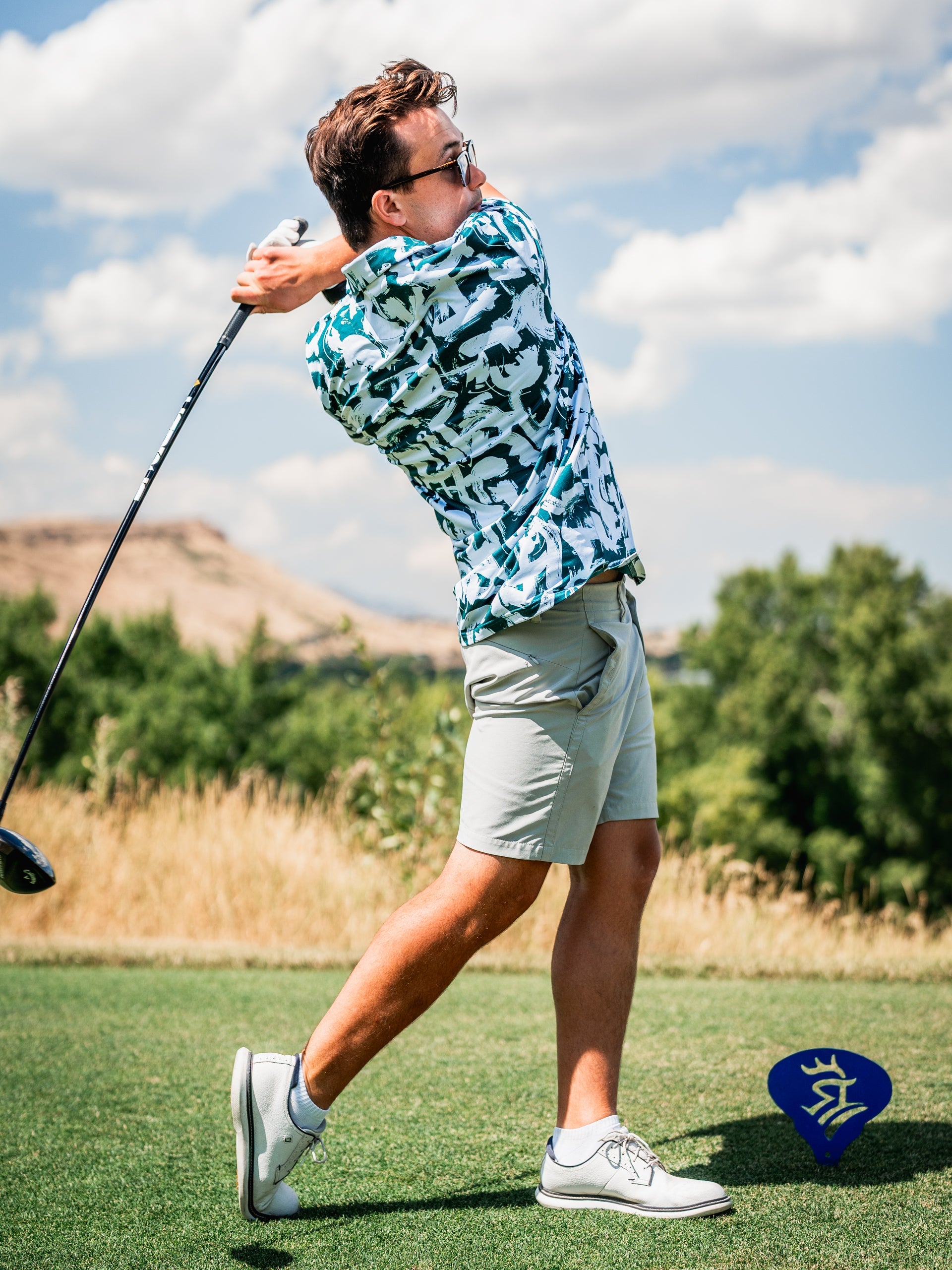 A golfer swings a club in a vibrant green paint strokes patterned shirt, paired with light shorts and stylish shoes.