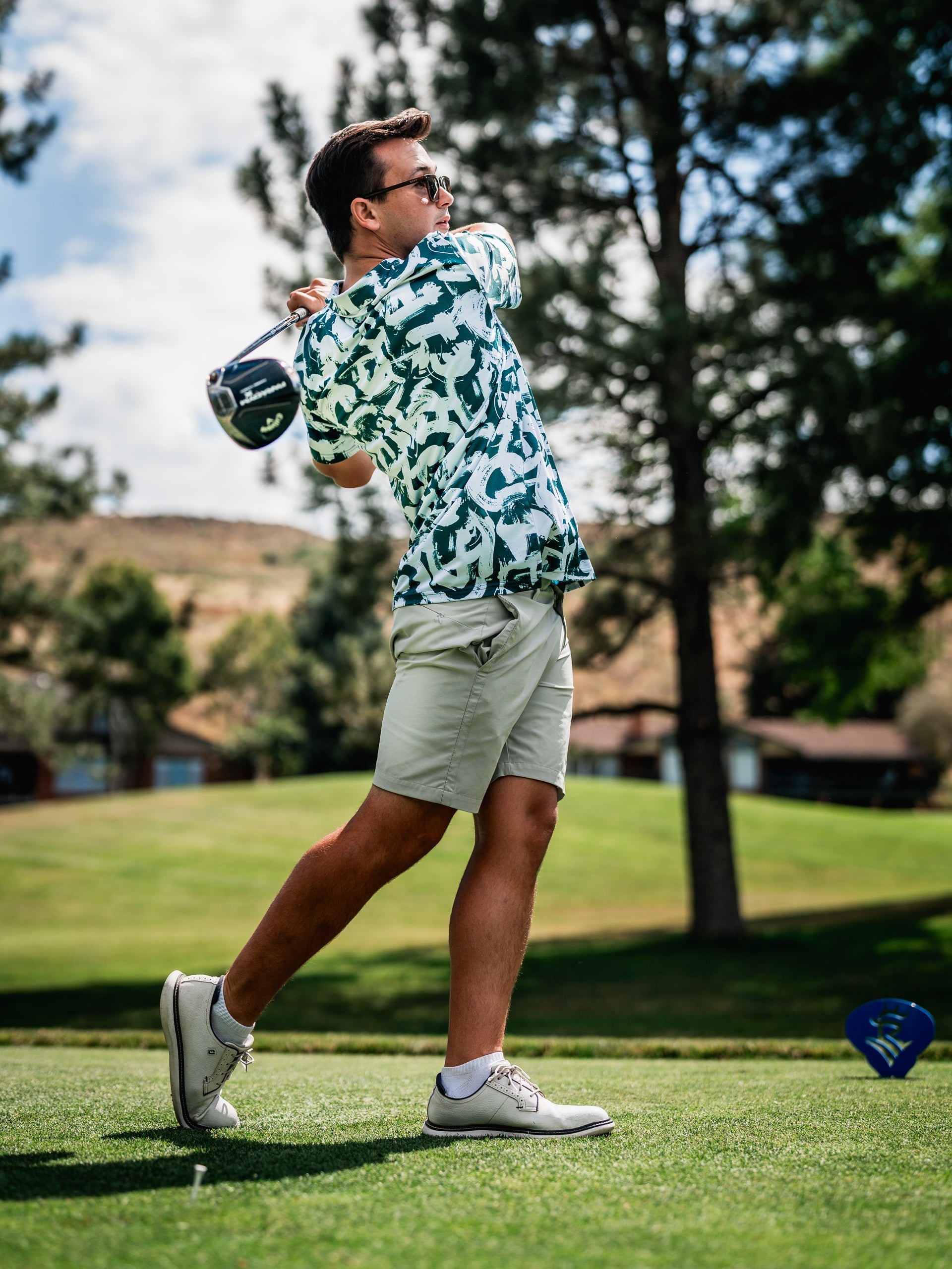 A golfer swings a club on the course, wearing a green and white patterned shirt and beige shorts, showcasing a stylish look.