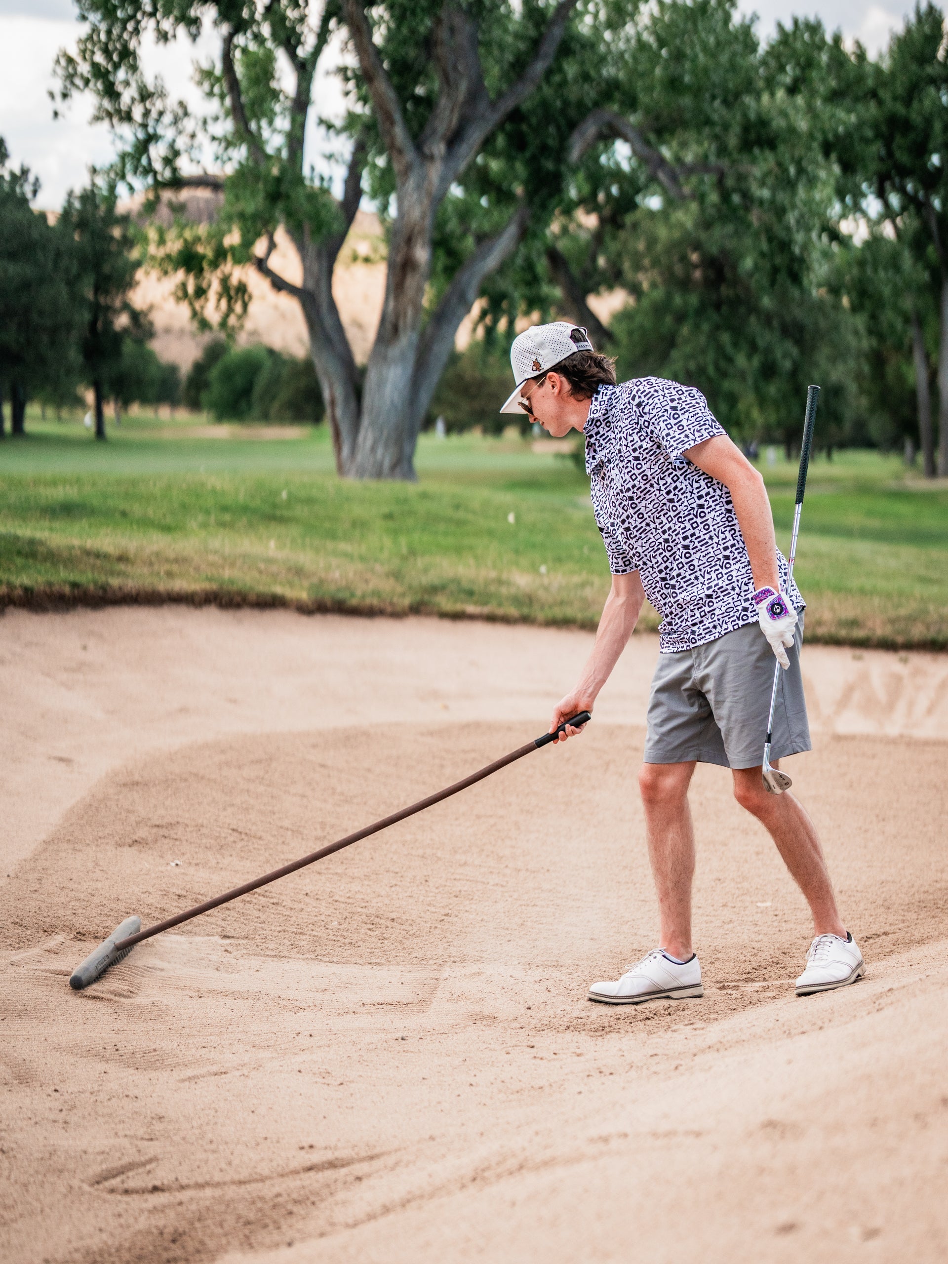Young man in a patterned shirt and white cap golfing from a sand trap on a lush course, holding a golf club and unique hea...