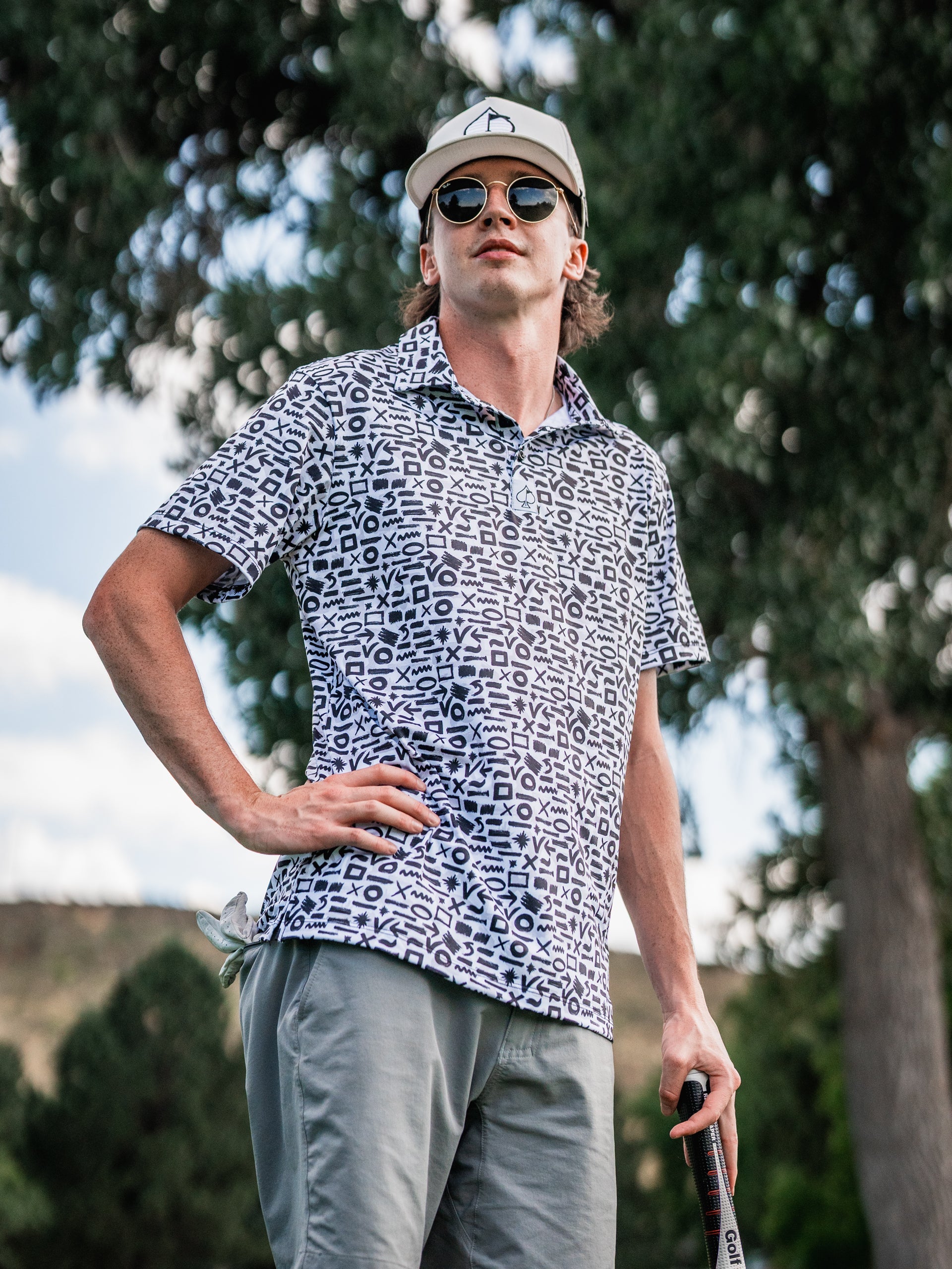 Male golfer in a black and white geometric patterned polo, wearing sunglasses and a white cap, holding a golf club outdoors.