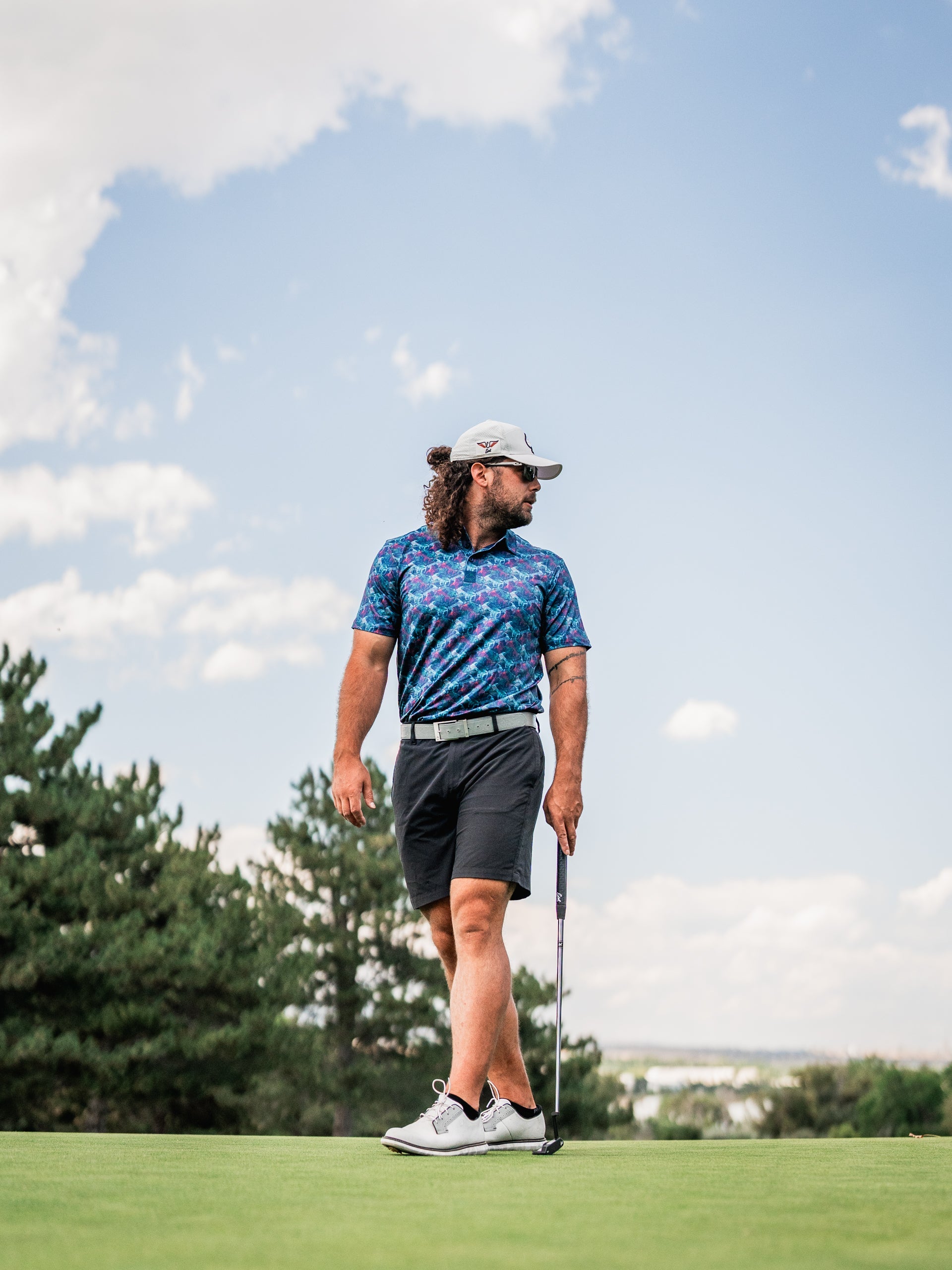 A golfer wearing a vibrant Swordfish Skelly polo shirt and shorts stands on the green, holding a putter under a blue sky.