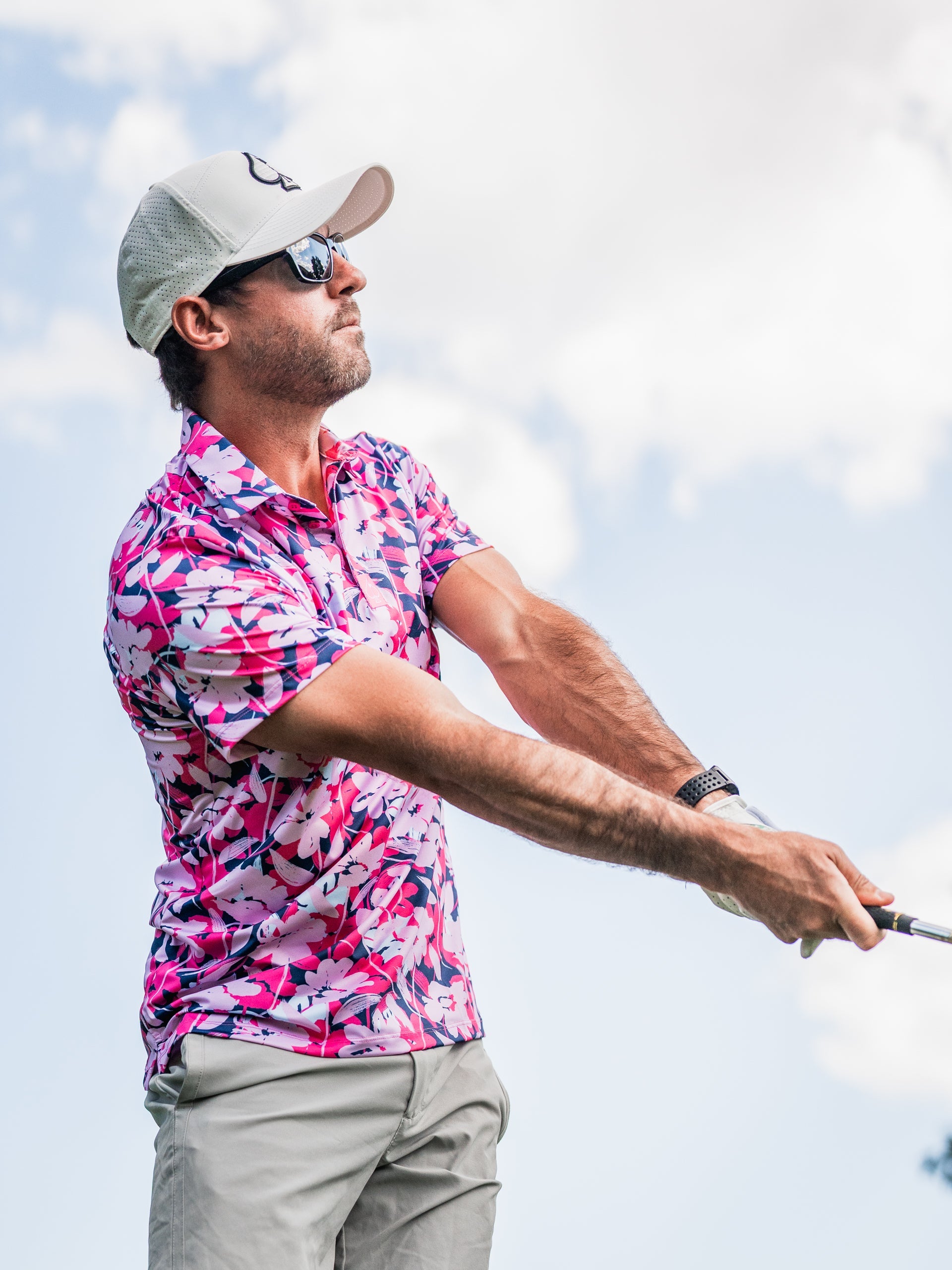 Man in pink floral golf shirt, beige cap, and sunglasses swinging a golf club outdoors against cloudy sky.