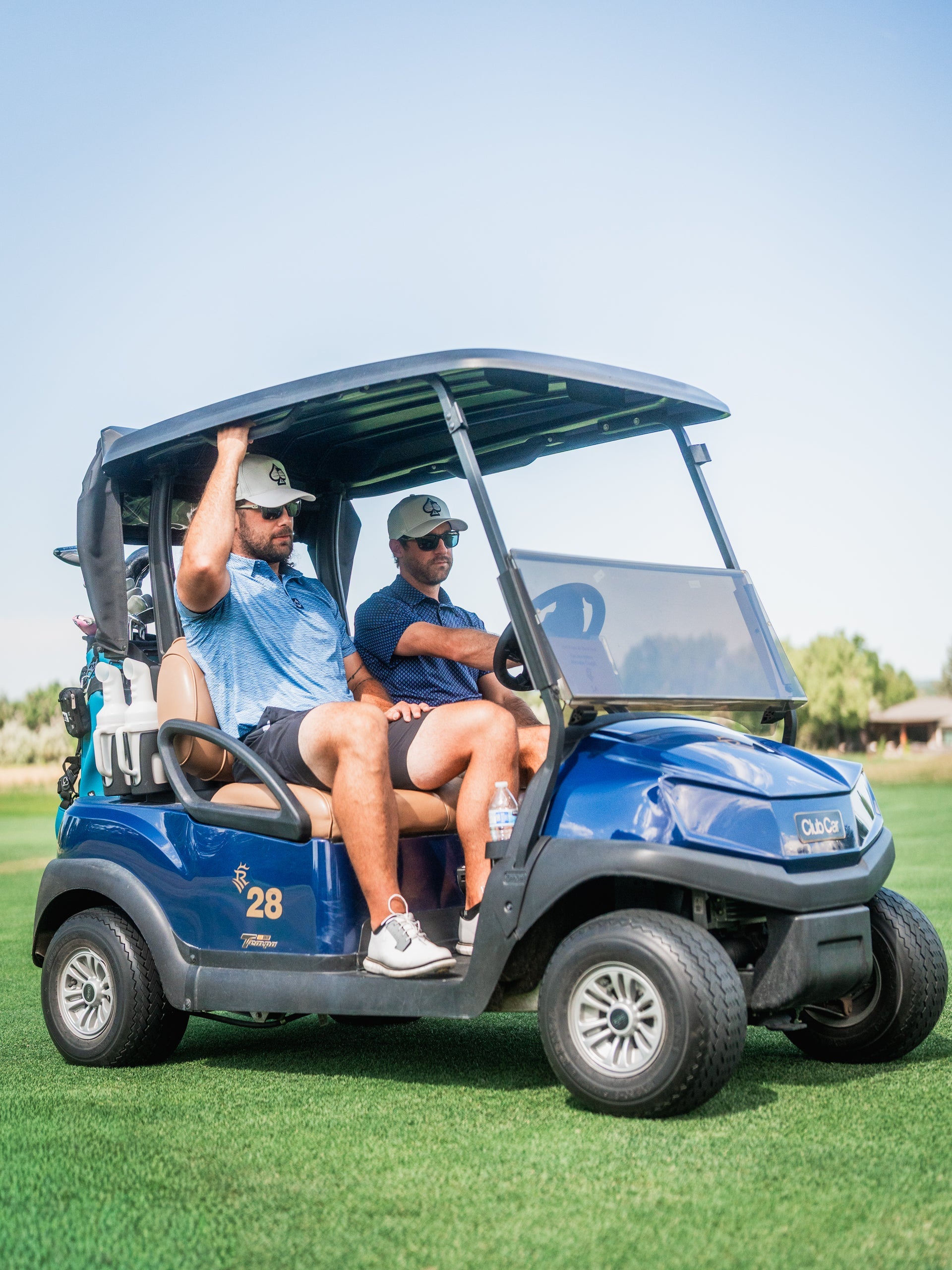 Two men in golf attire sit in a blue golf cart on the course, under a clear sky, with bold, sporty style accessories.