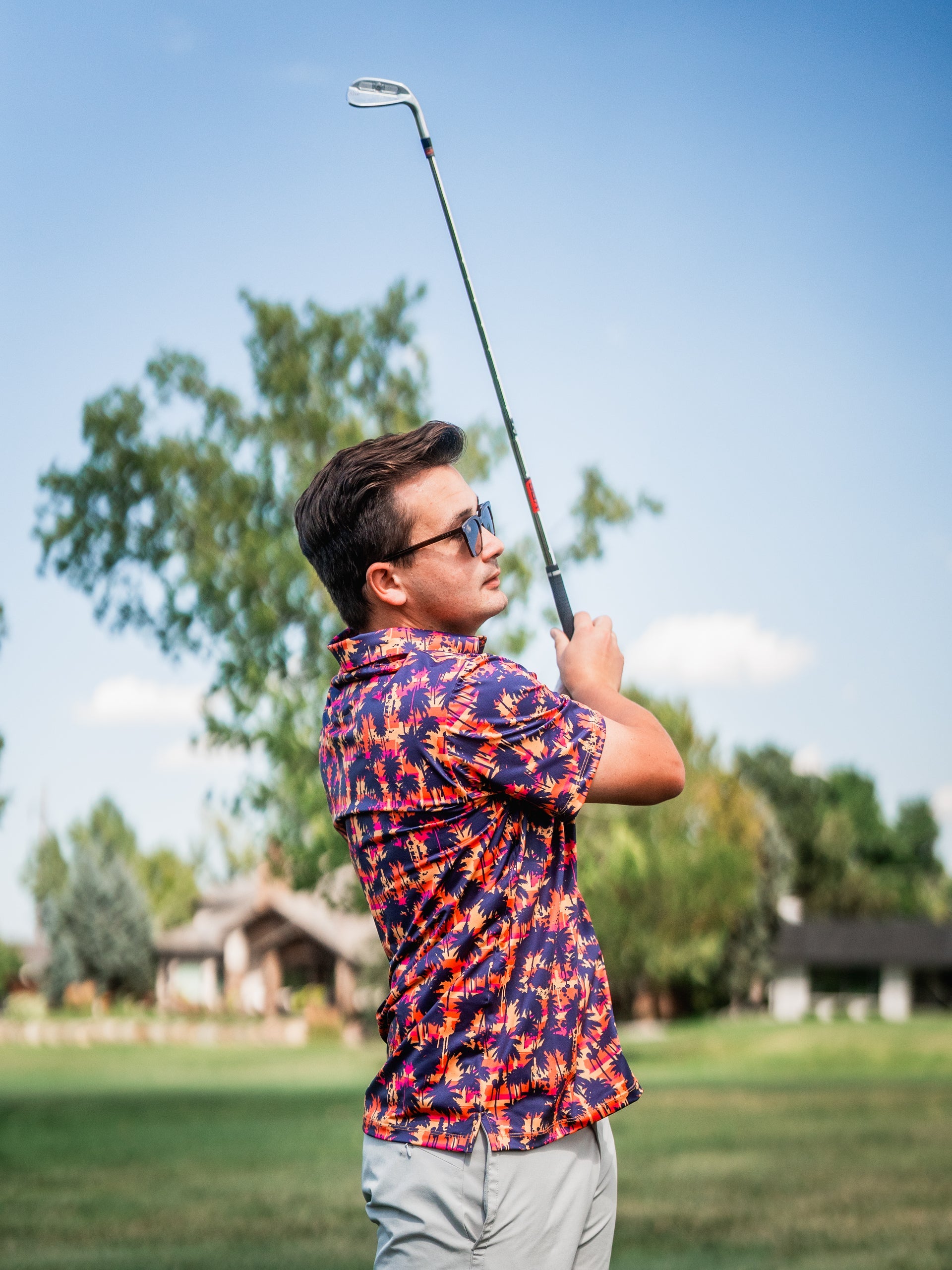 Young man in colorful tropical golf shirt with sunglasses, swinging golf club on course under blue sky.