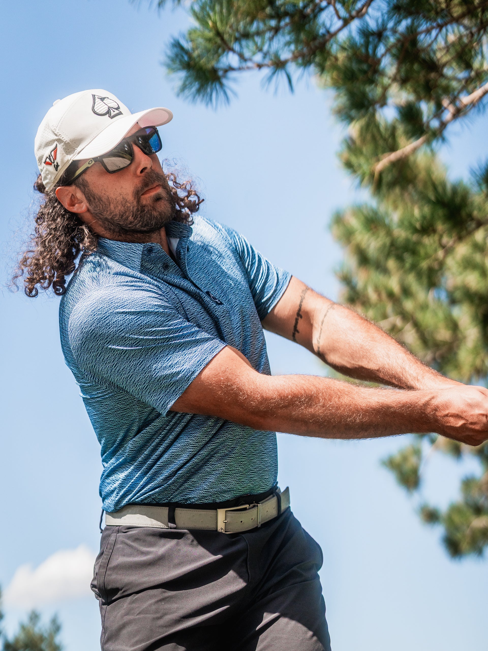 A golfer swings a club while wearing a blue patterned polo shirt, sunglasses, and a cap, set against a sunny backdrop.