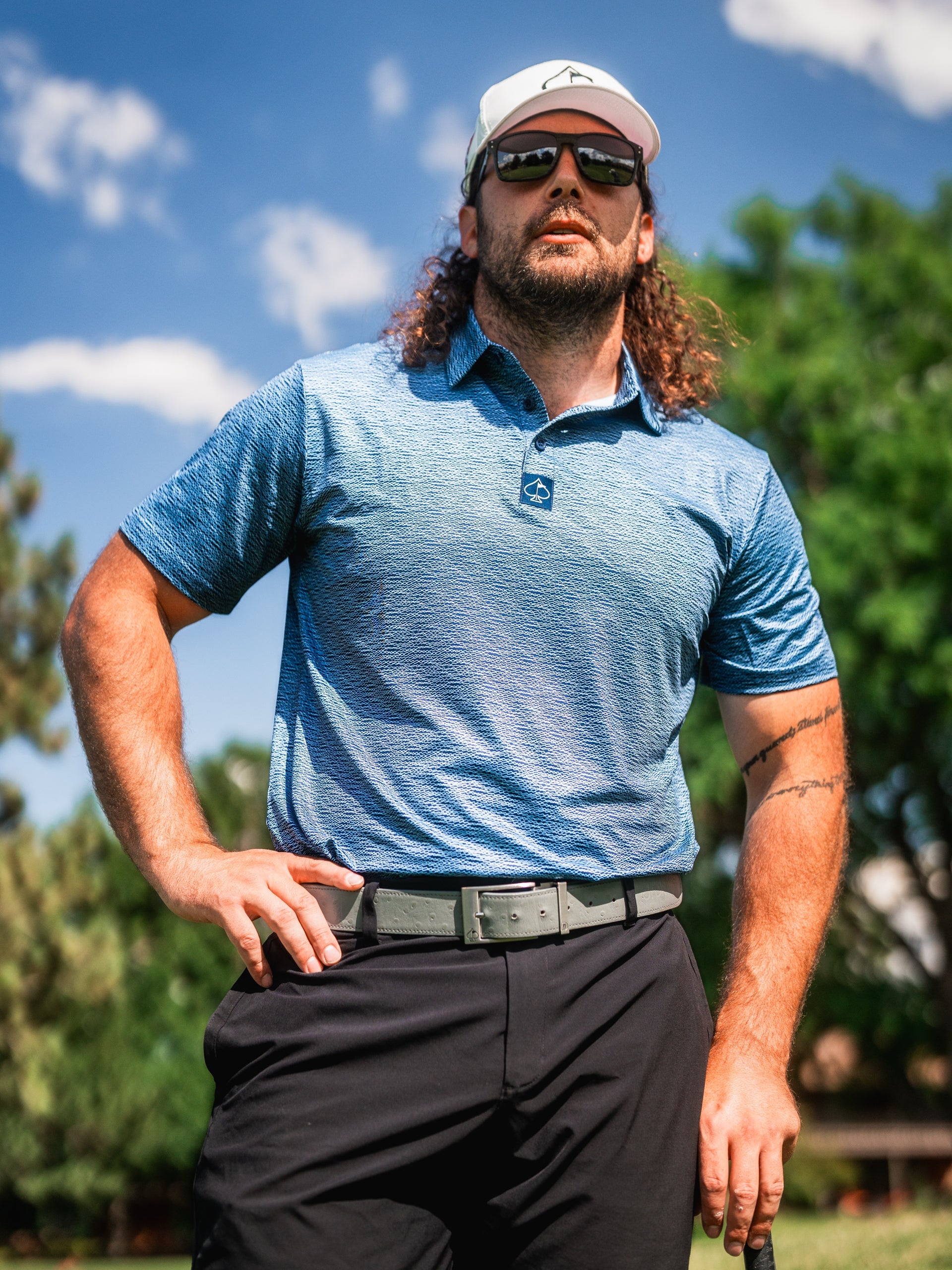 Man wearing a blue golf polo with a squiggle pattern, standing confidently on a golf course under a clear sky.