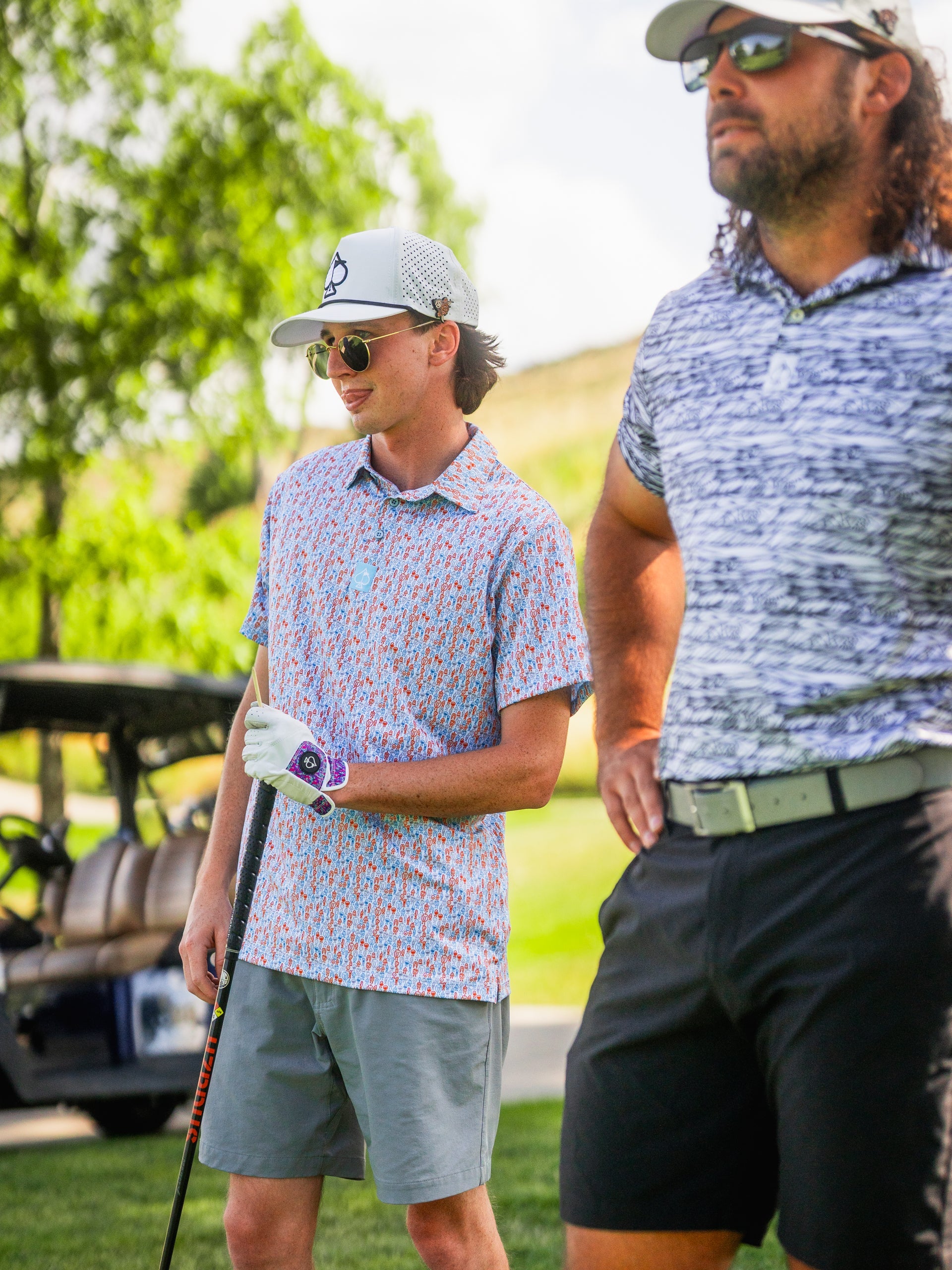 Young man in a colorful patterned golf shirt, gray shorts, and a white cap with sunglasses, holding a golf club on a sunny...