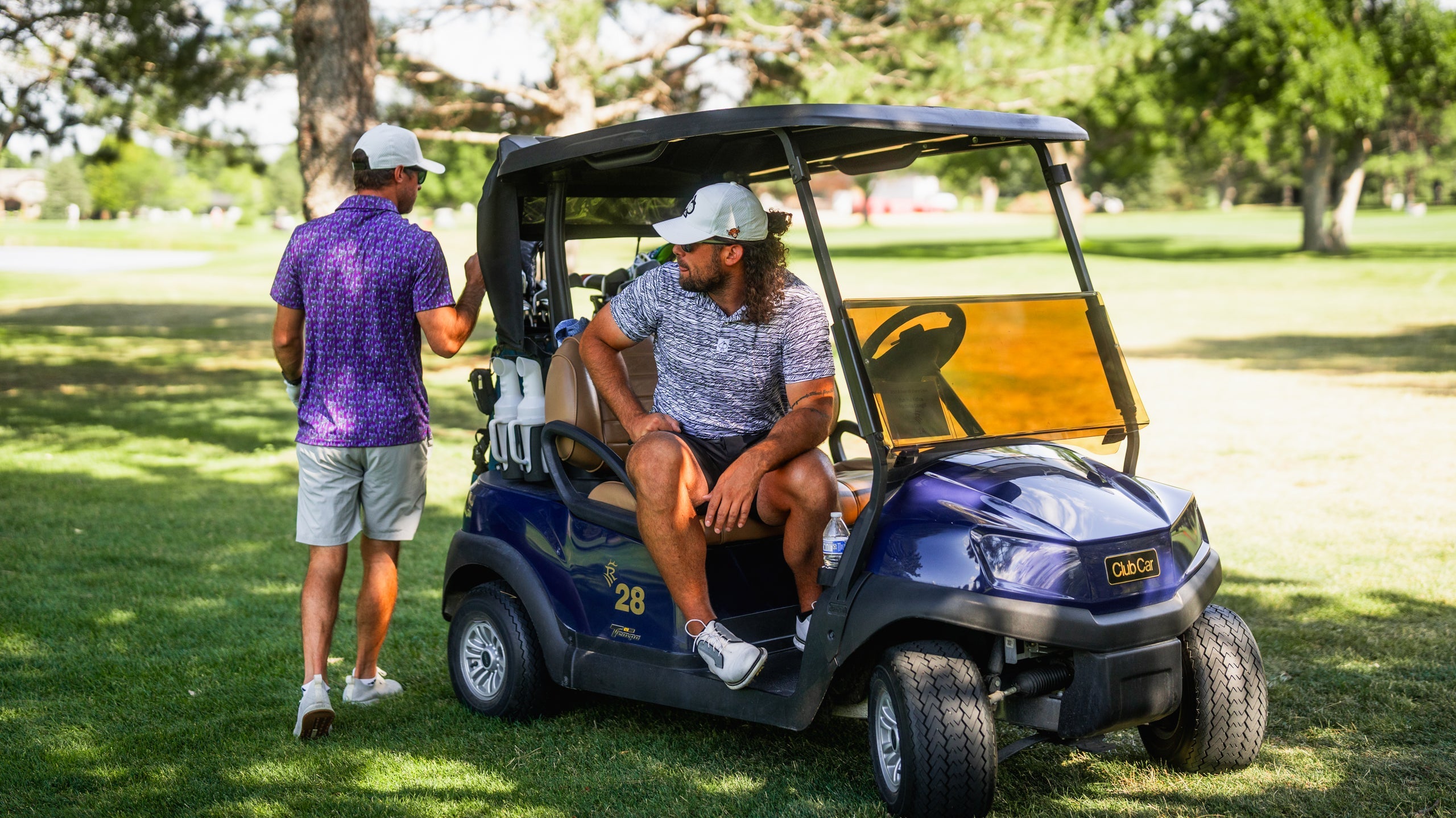 A golfer in a Fragment Black shirt relaxes on a golf cart while another golfer stands nearby, both enjoying a sunny day on the course.